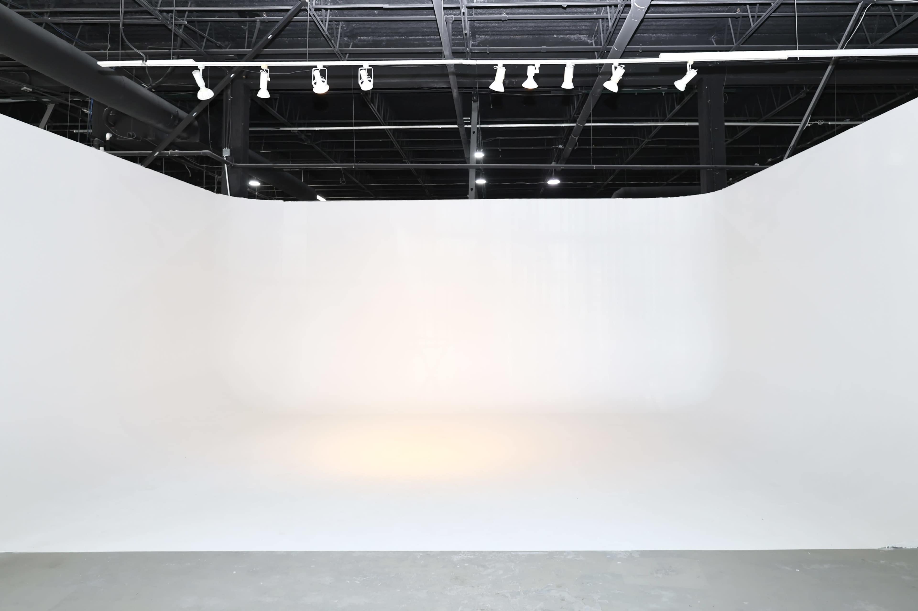 The image shows a spacious, minimally furnished white photography studio with a seamless backdrop and overhead lighting.