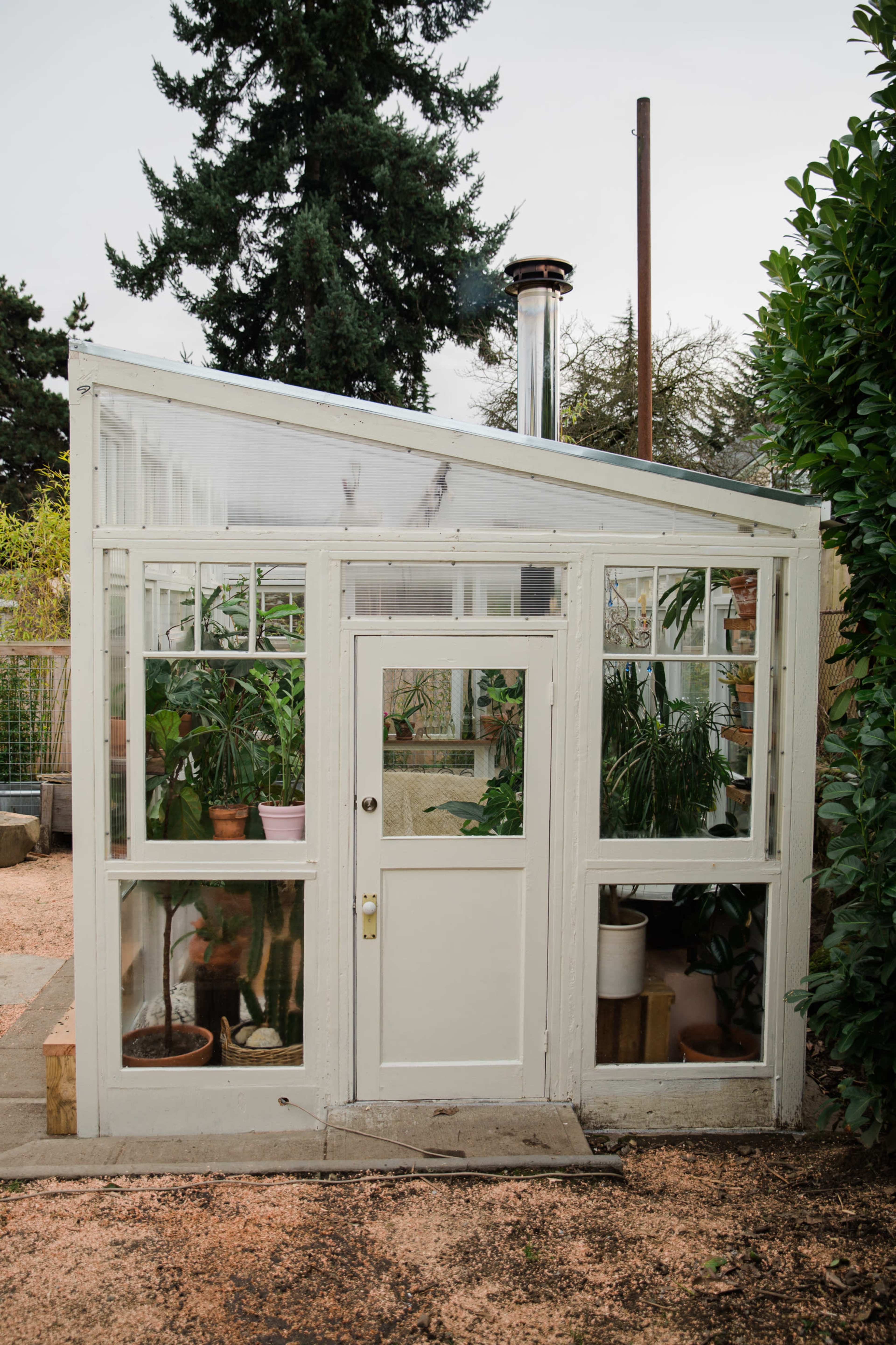 A small greenhouse with glass walls and a door is filled with potted plants and situated in a landscaped garden.