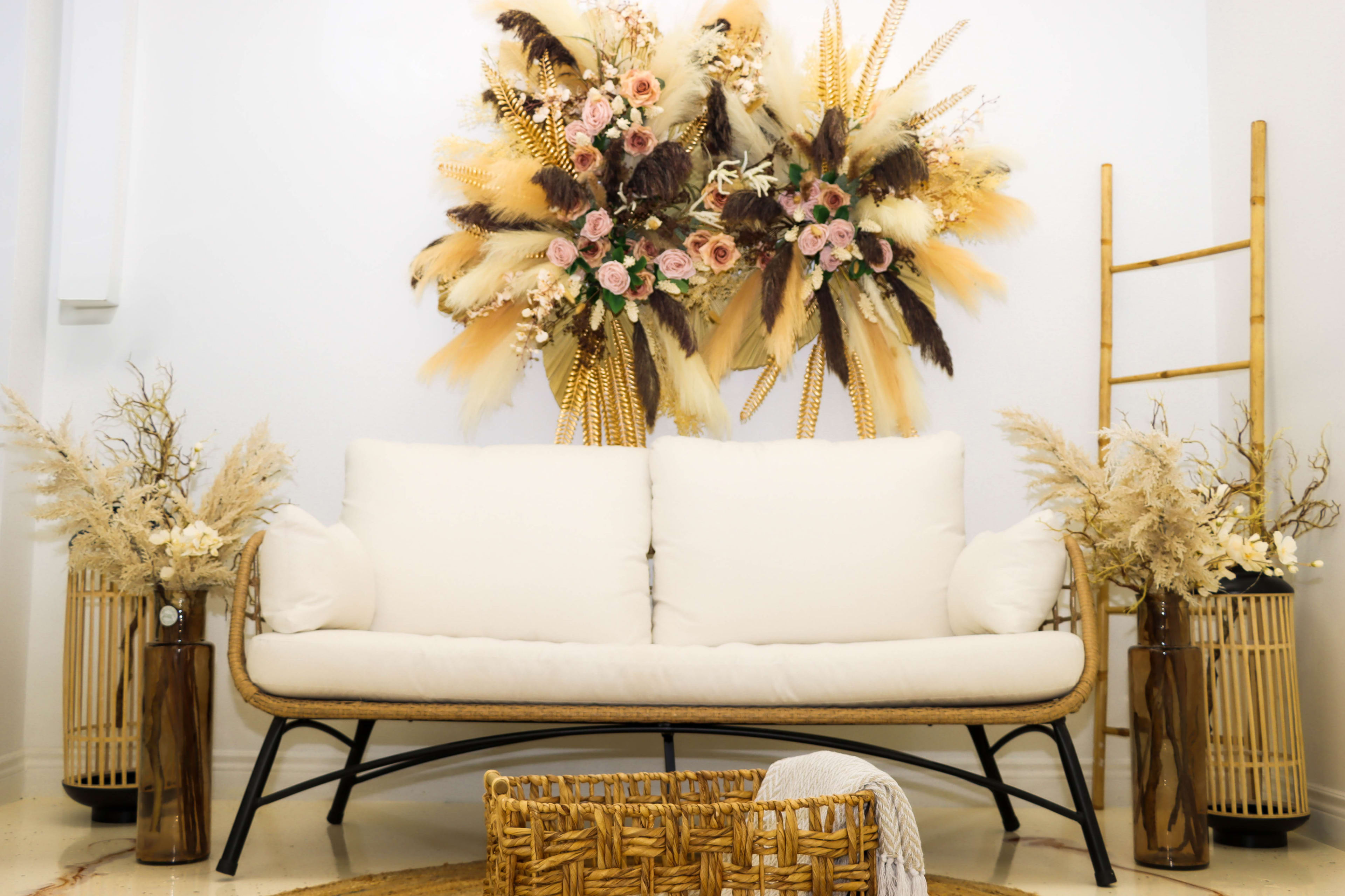 A white sofa is positioned in front of a decorative floral arrangement made of dried plants and flowers, with decorative vases and a woven basket nearby.