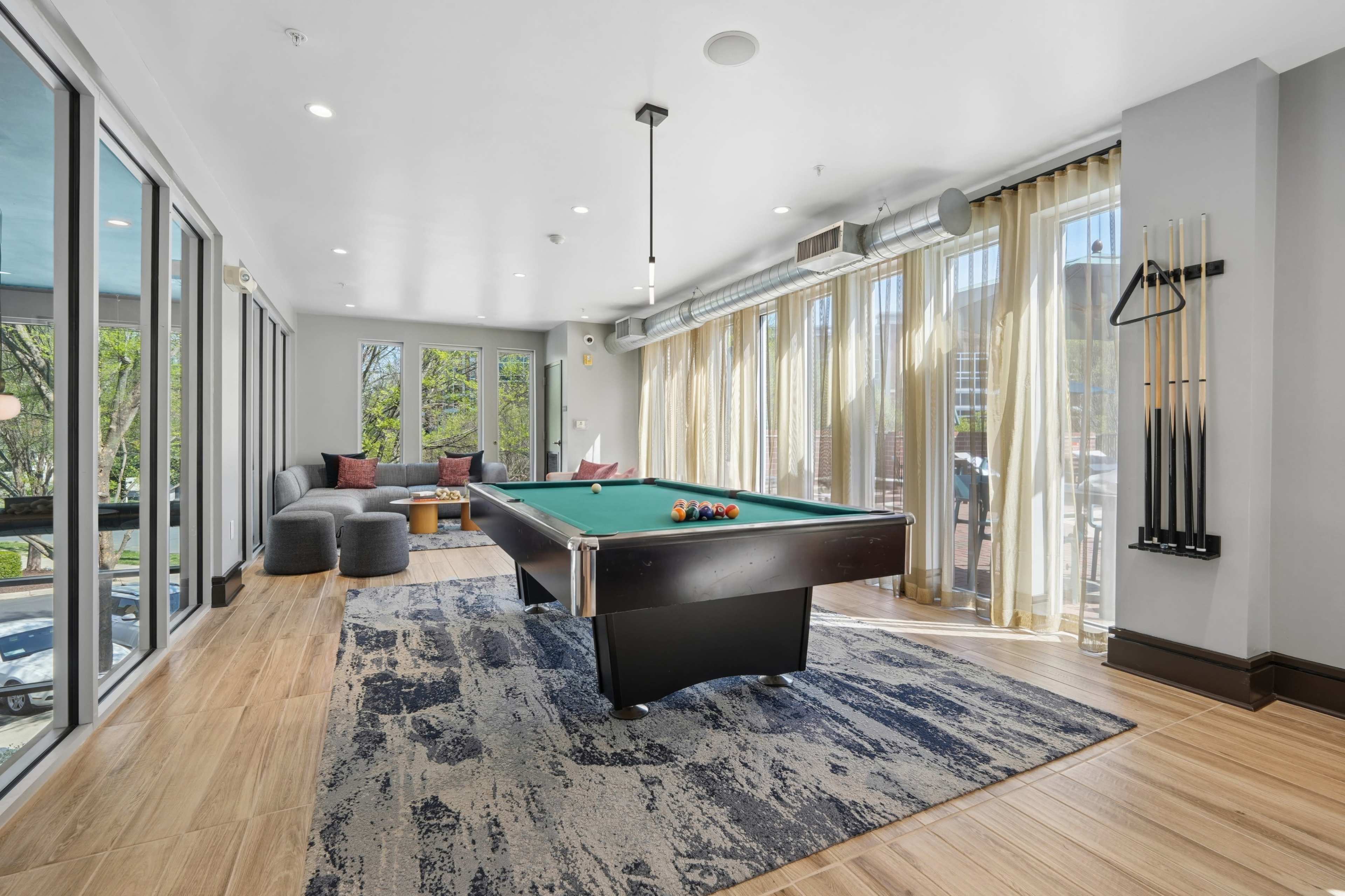 The image shows a well-lit room with large windows, featuring a billiards table, two sofas, and a decorative rug.