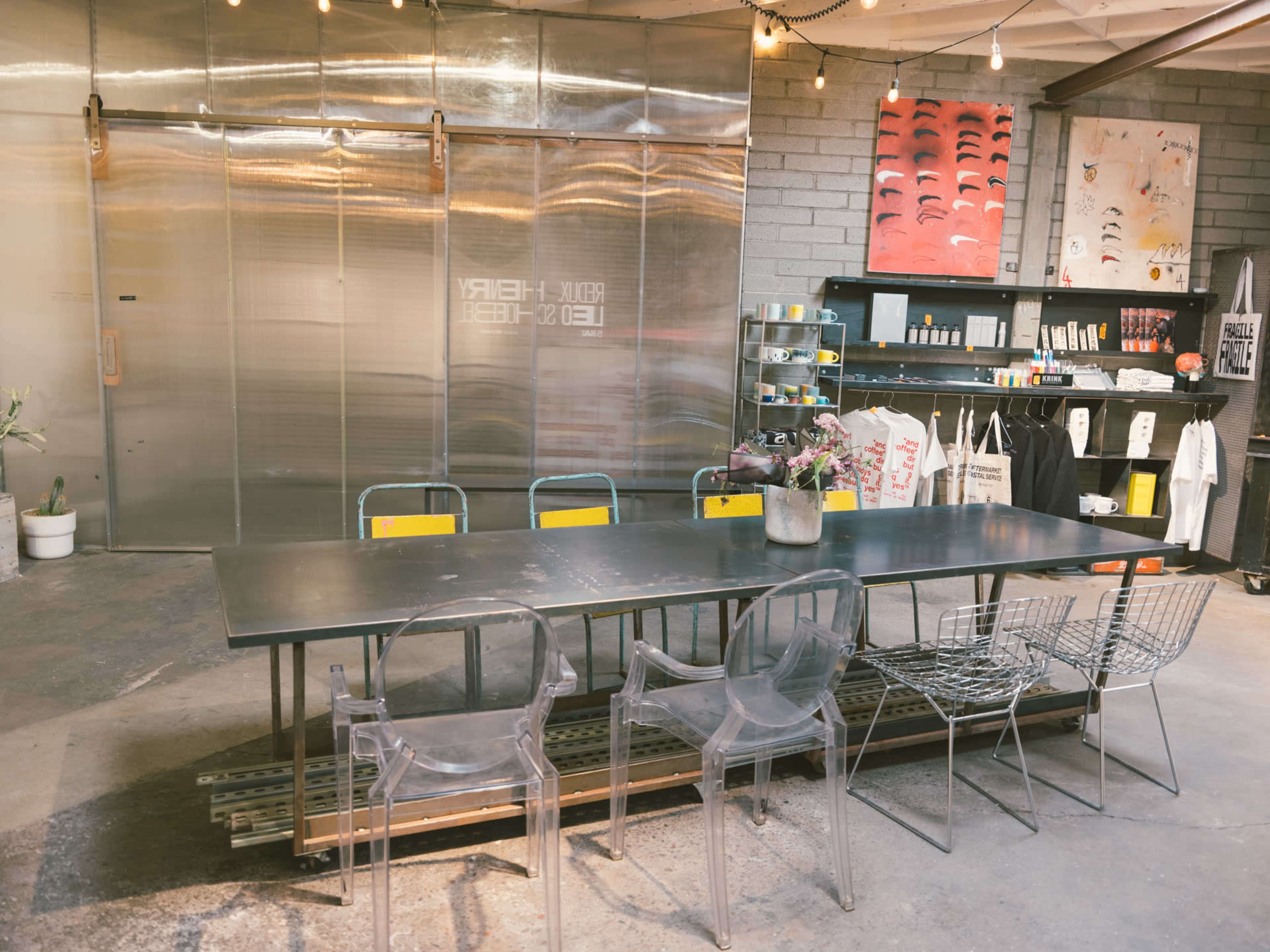 A long, dark table with transparent chairs is set up in an industrial-style room featuring metal sliding doors and various artworks on the walls.
