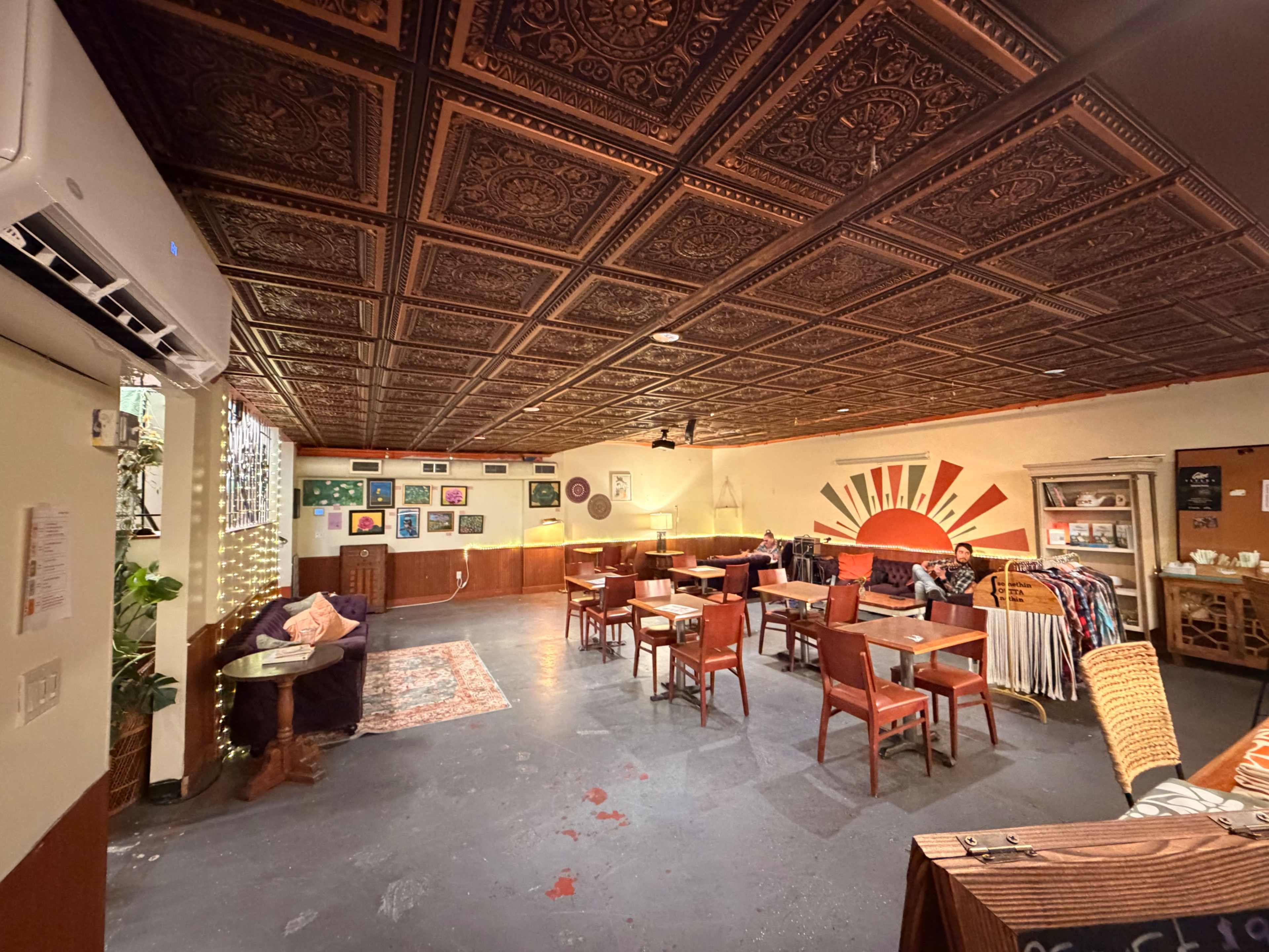 The image shows a spacious café or lounge with a patterned ceiling, various tables and chairs, and colorful artwork displayed on the walls.