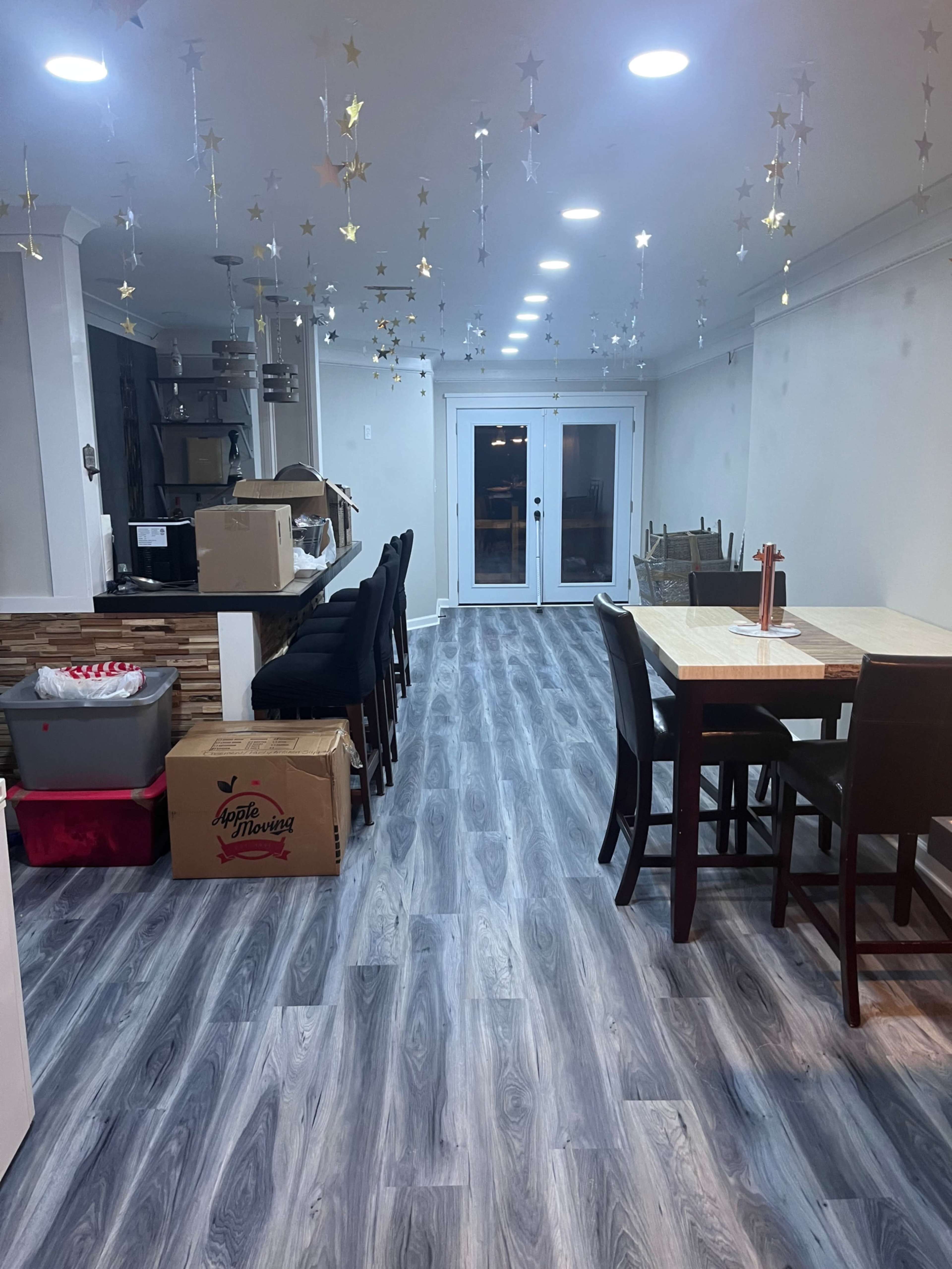 A partially furnished interior space with a dining table and chairs, a counter with bar stools, and boxes stacked along the walls, all beneath a ceiling decorated with hanging stars.