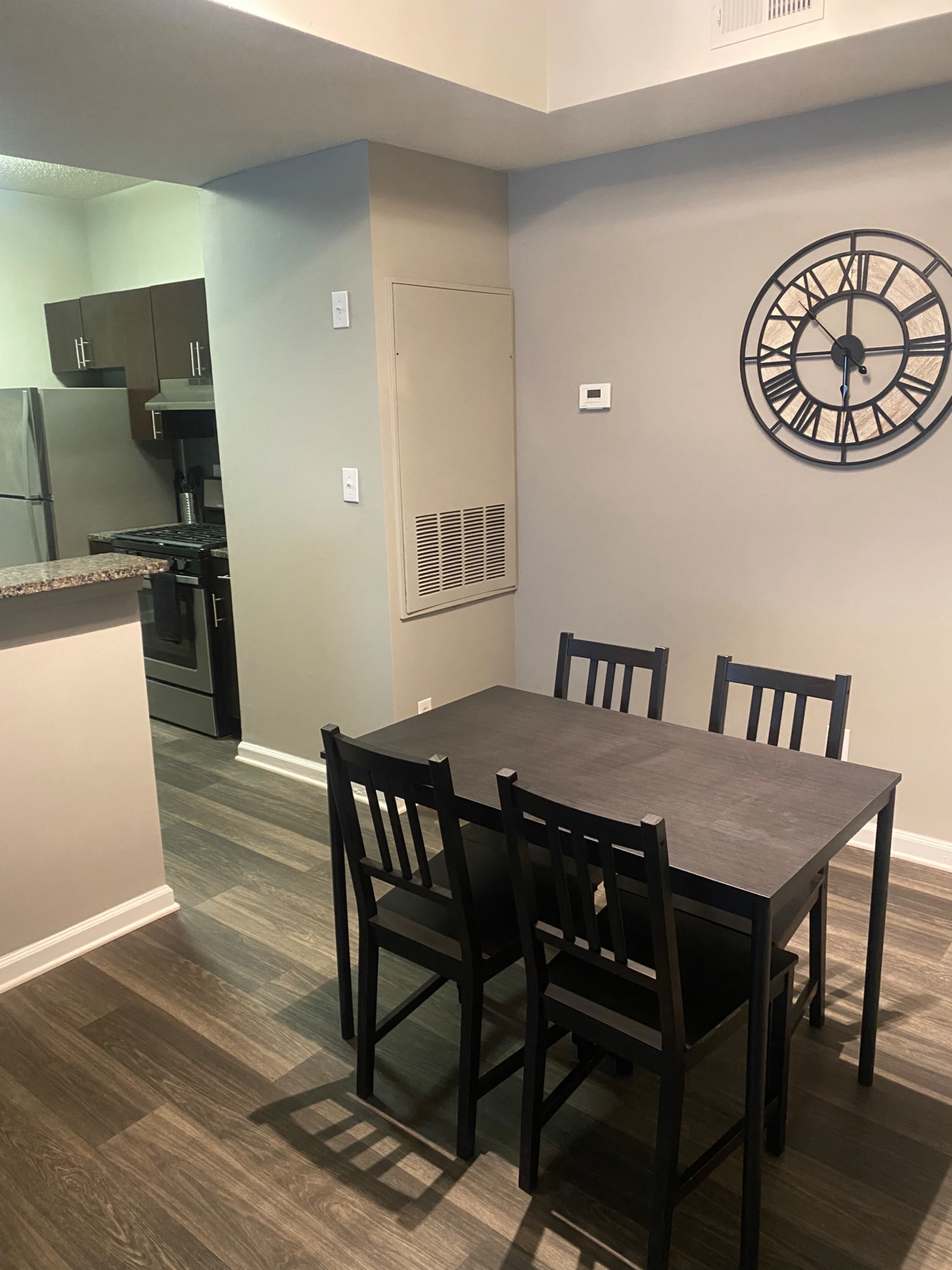 A small dining area features a dark table with four chairs, adjacent to a kitchen with stainless steel appliances and a wall clock.