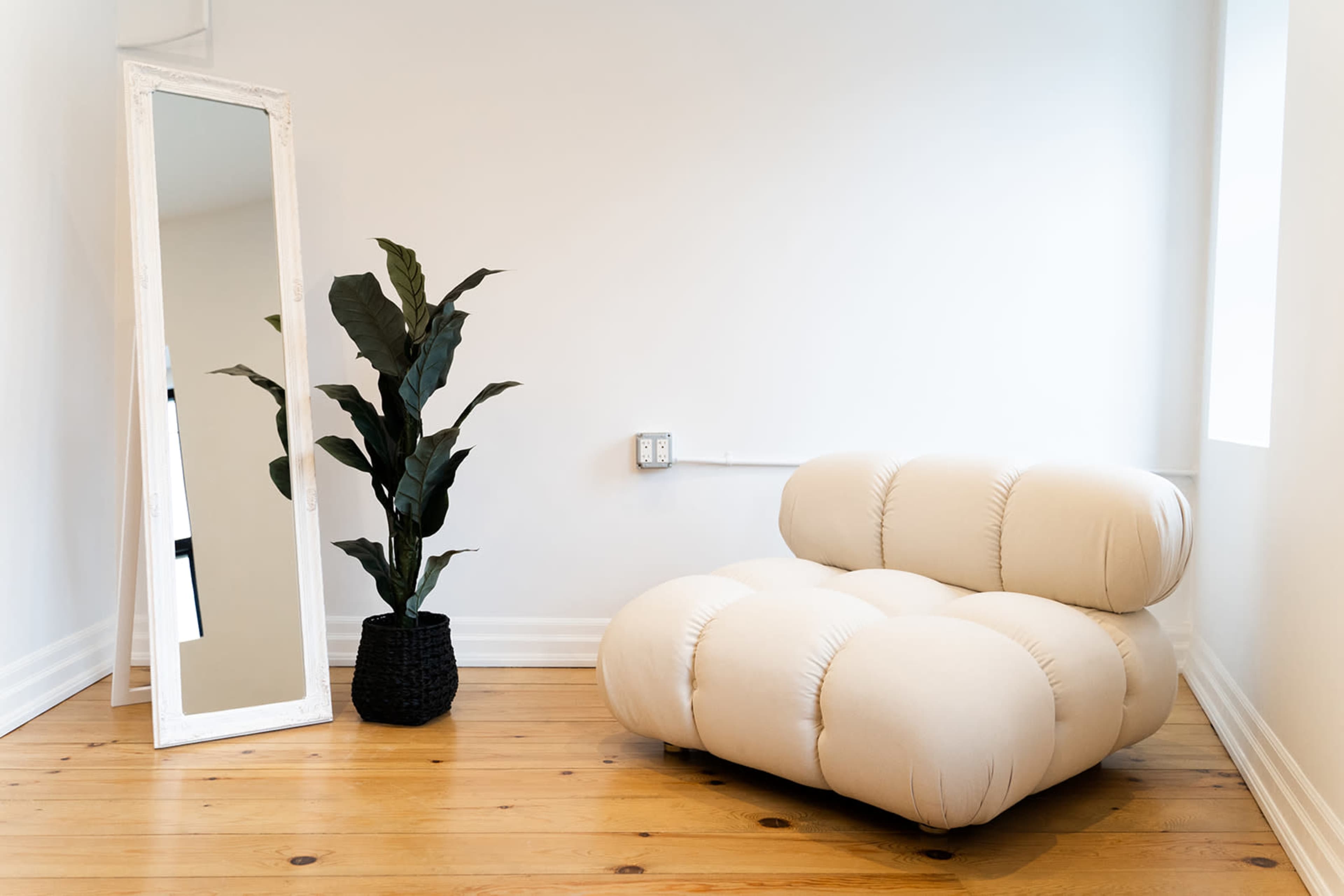 A minimalist room features a beige upholstered chair, a tall mirror, and a potted plant in a black container against a light-colored wall.