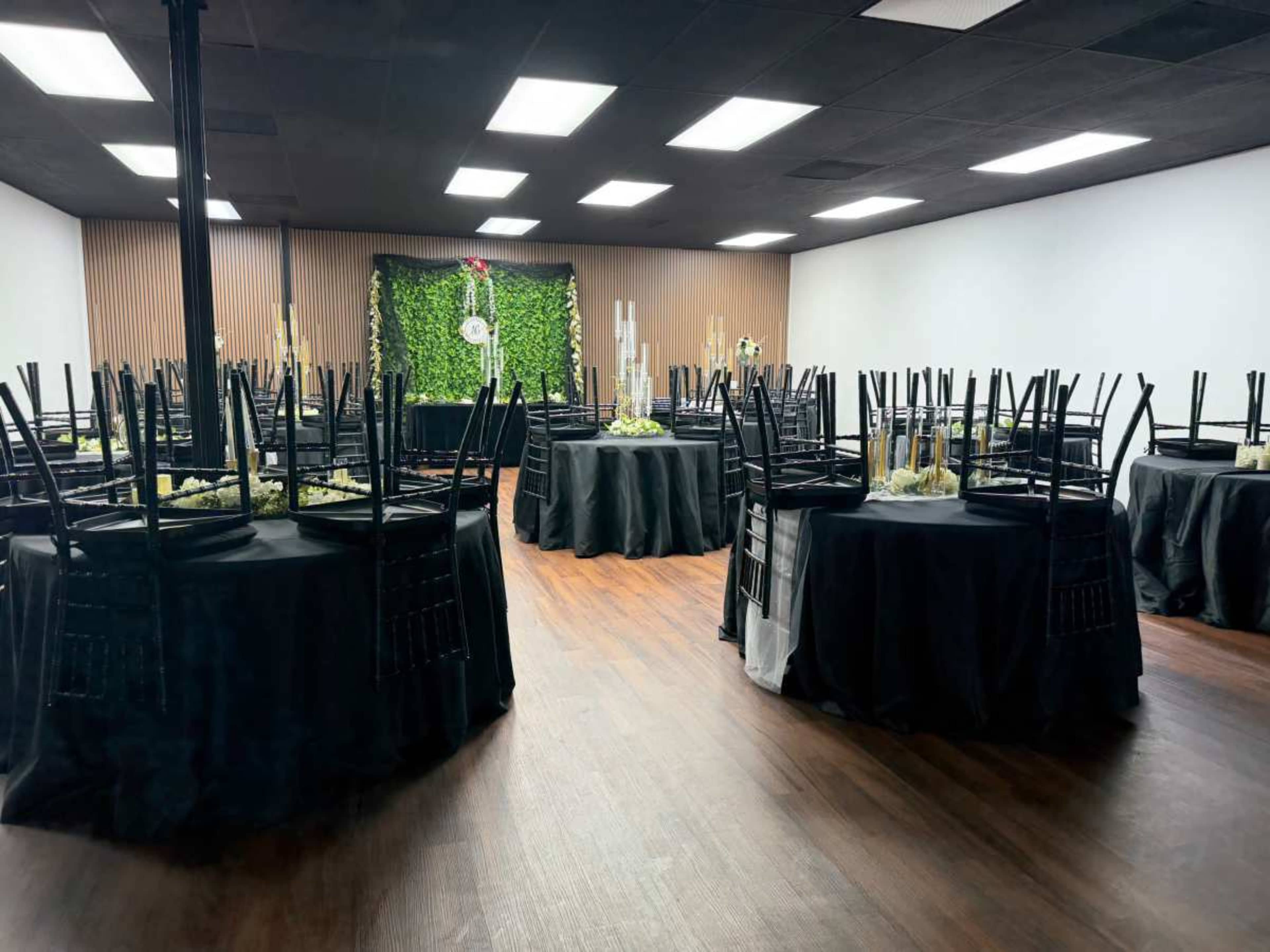 A banquet hall features several round tables covered with black tablecloths, each adorned with floral centerpieces, and chairs stacked neatly on top.
