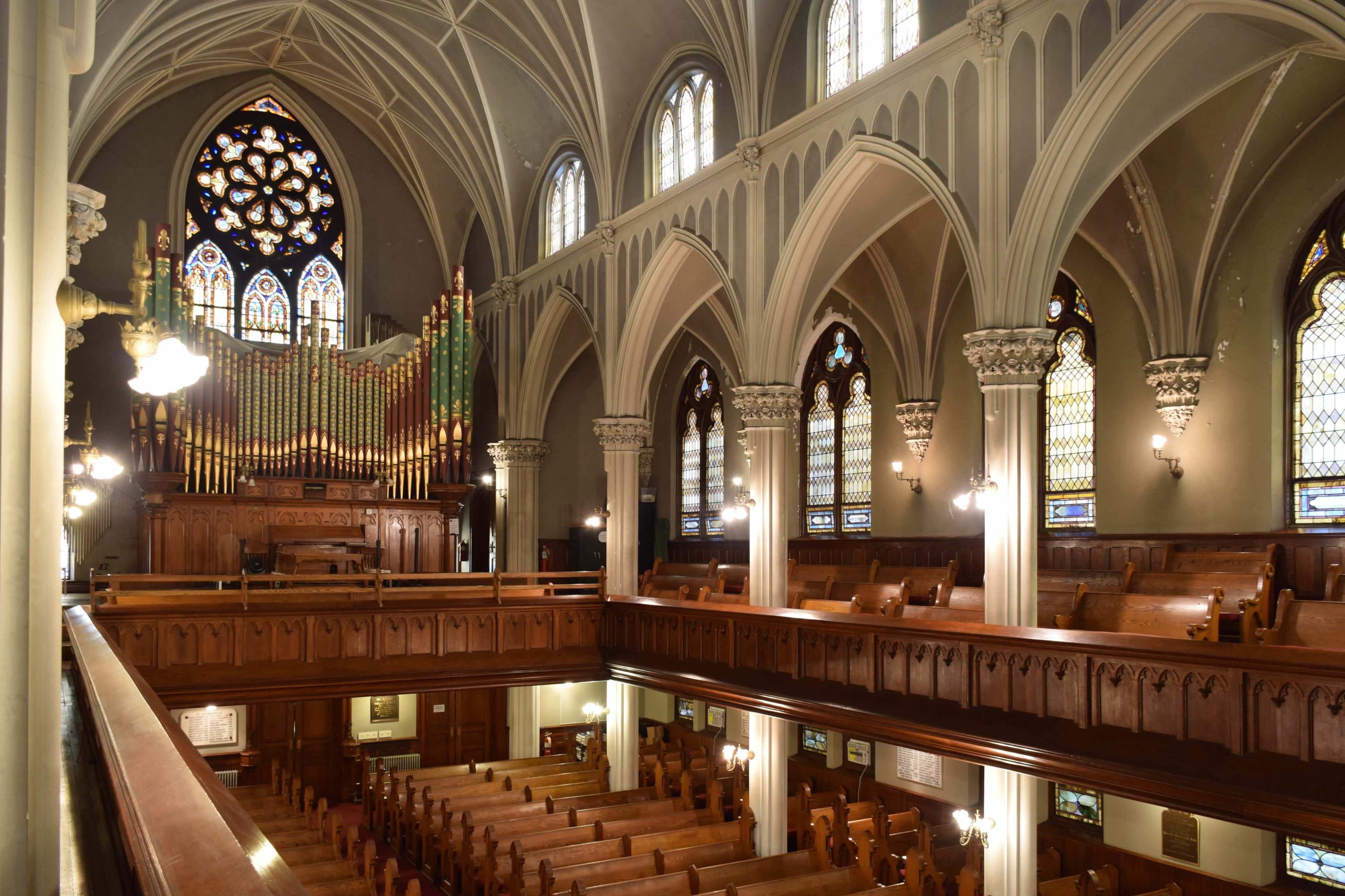 The interior of a Gothic-style church features high vaulted ceilings, a large organ at the back, and rows of wooden pews facing the altar.