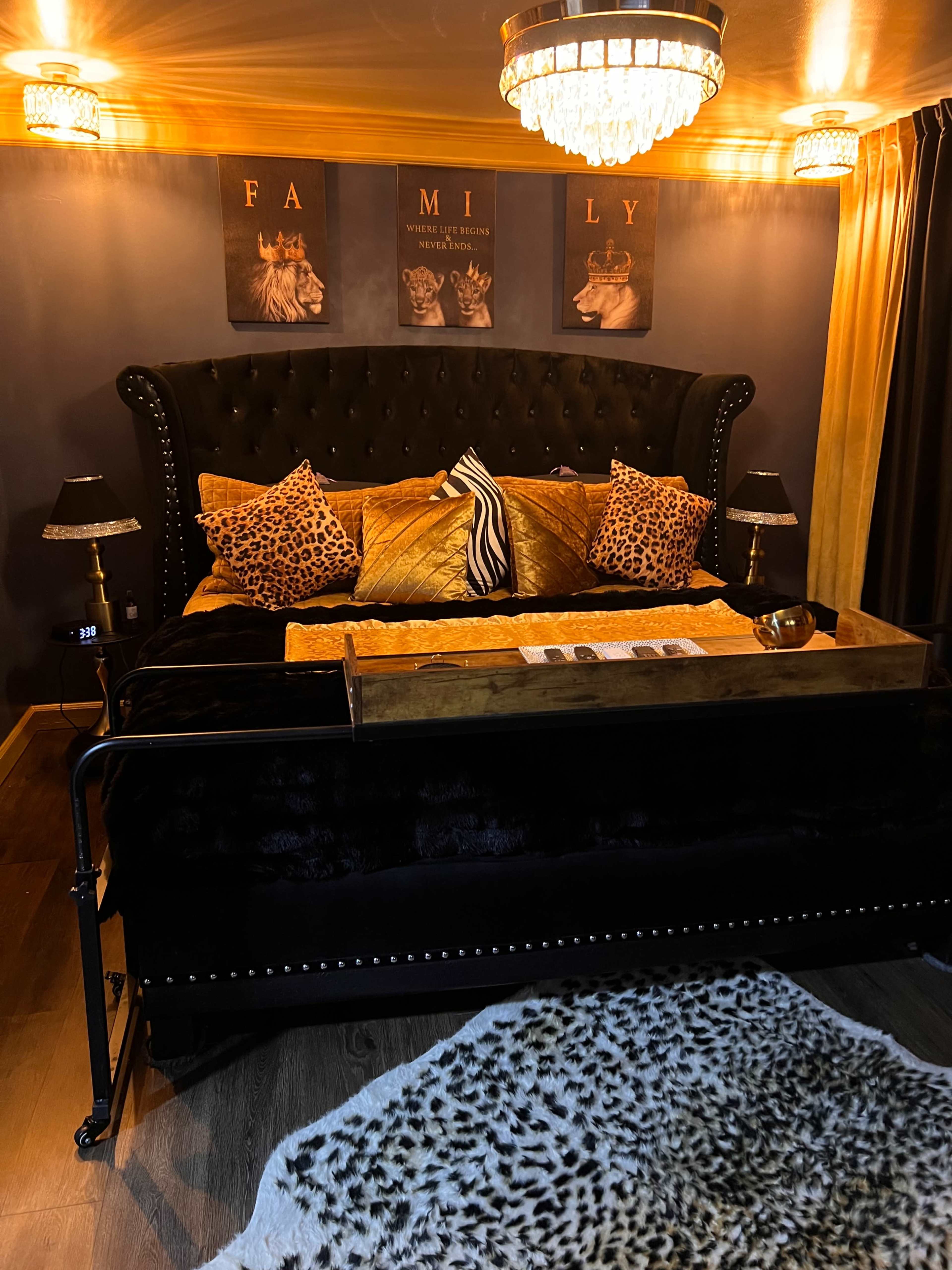 The image shows a luxurious bedroom featuring a large black upholstered bed with decorative pillows, a wooden tabletop, and a large area rug on the floor.
