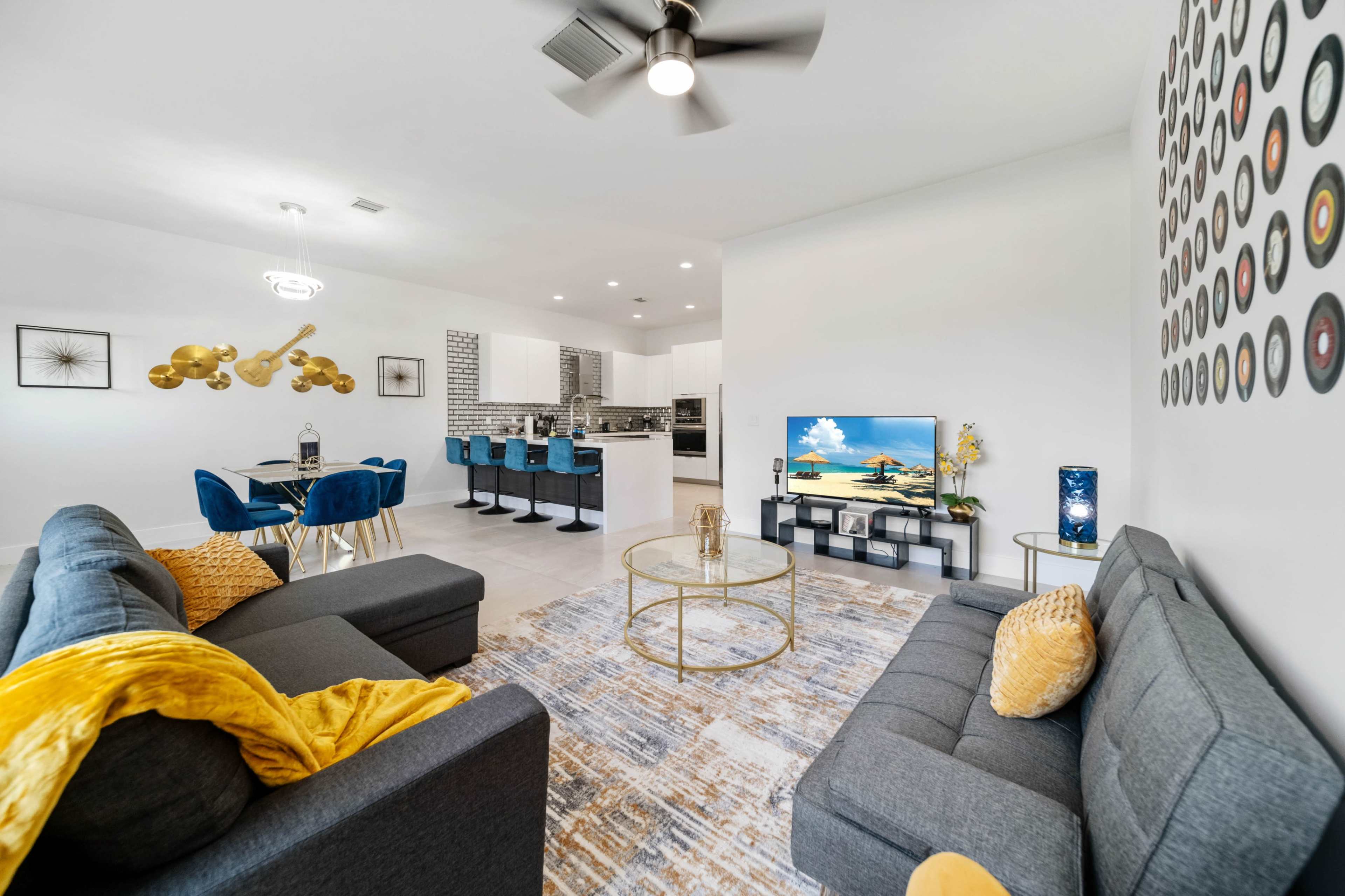 The image shows a modern living room with two gray sofas, a glass coffee table, and a television, adjacent to a dining area featuring blue chairs and a kitchen with white cabinetry.