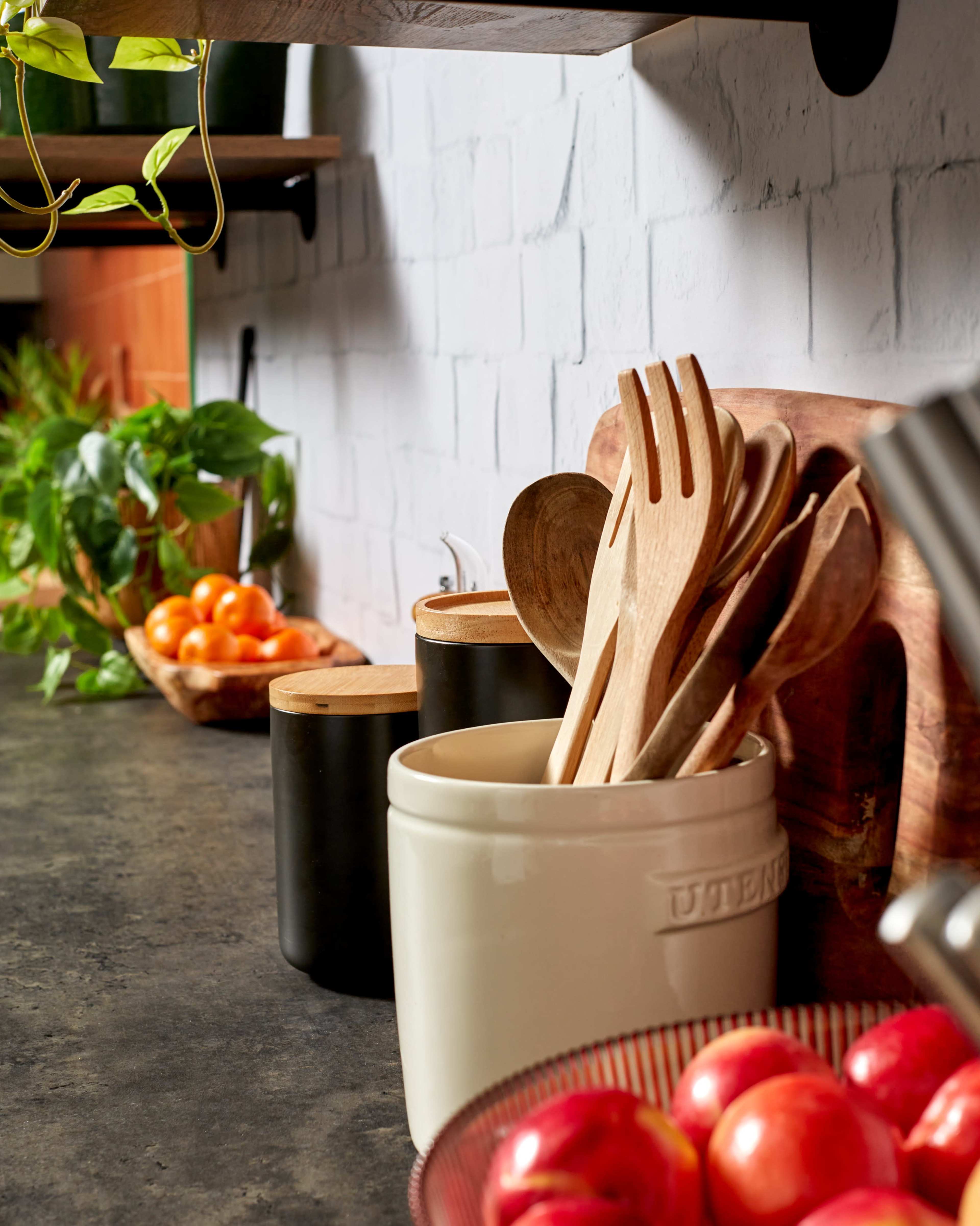 A countertop displays various cooking utensils in jars, a bowl of apples, and a small plant, set against a textured wall.