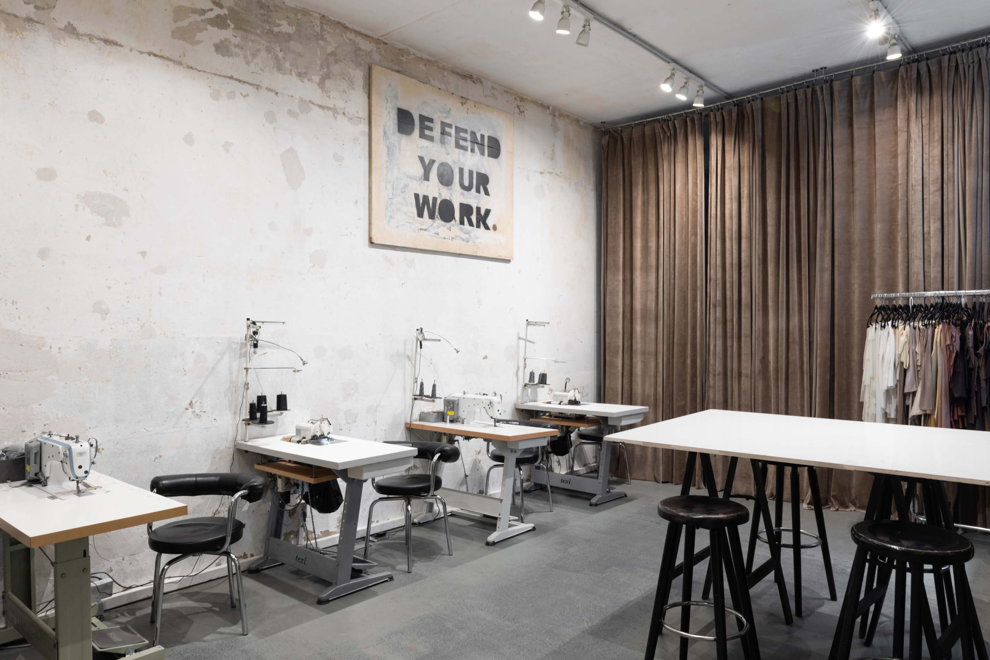 A sewing workshop with multiple sewing machines against a textured wall and a large banner overhead reading "DEFEND YOUR WORK."