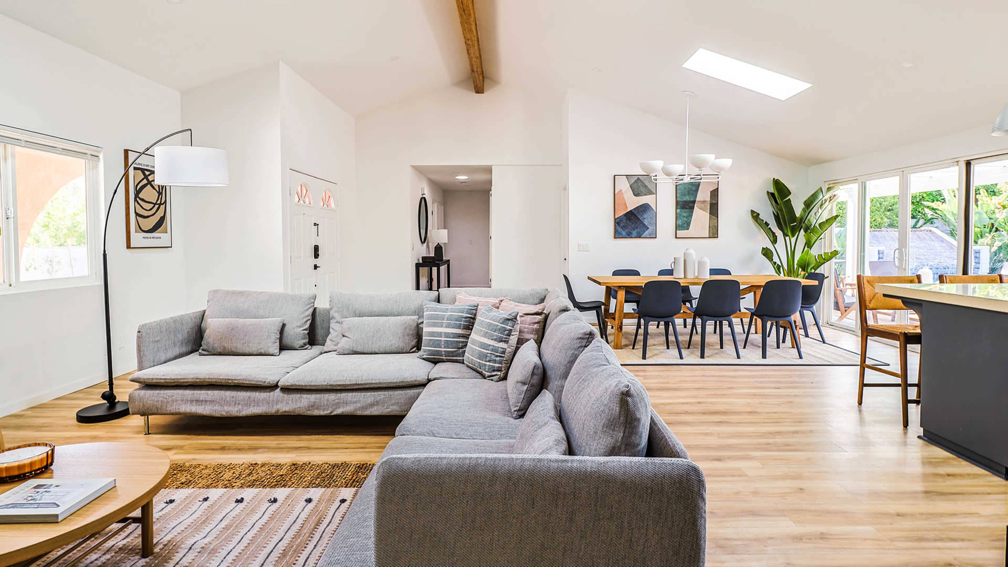 A modern, open-concept living room and dining area with a gray sofa, a wooden dining table, and large windows letting in natural light.