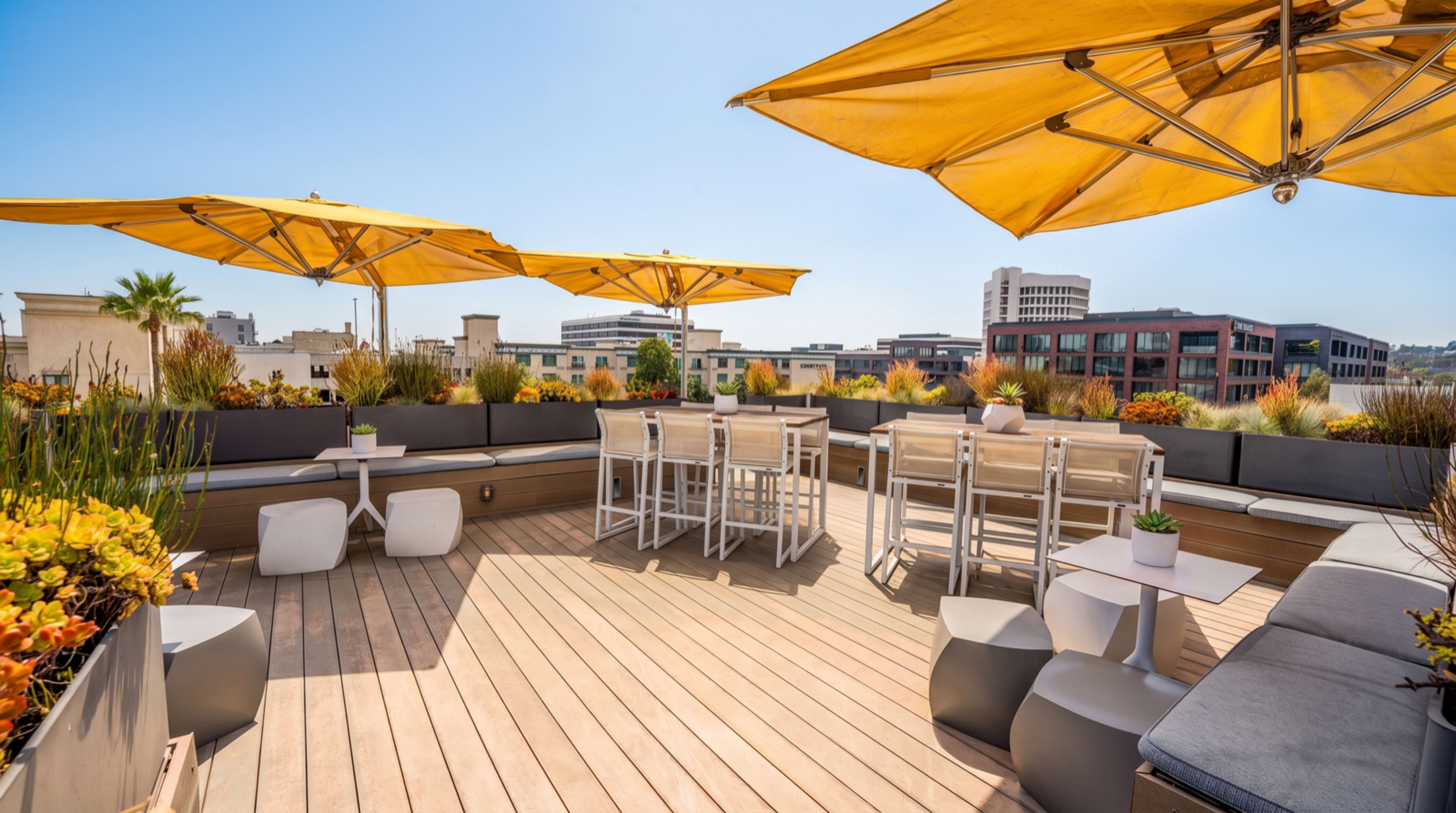 The image shows a rooftop terrace with wooden decking, white tables and chairs, and yellow umbrellas, surrounded by plants and buildings in the background under a clear blue sky.
