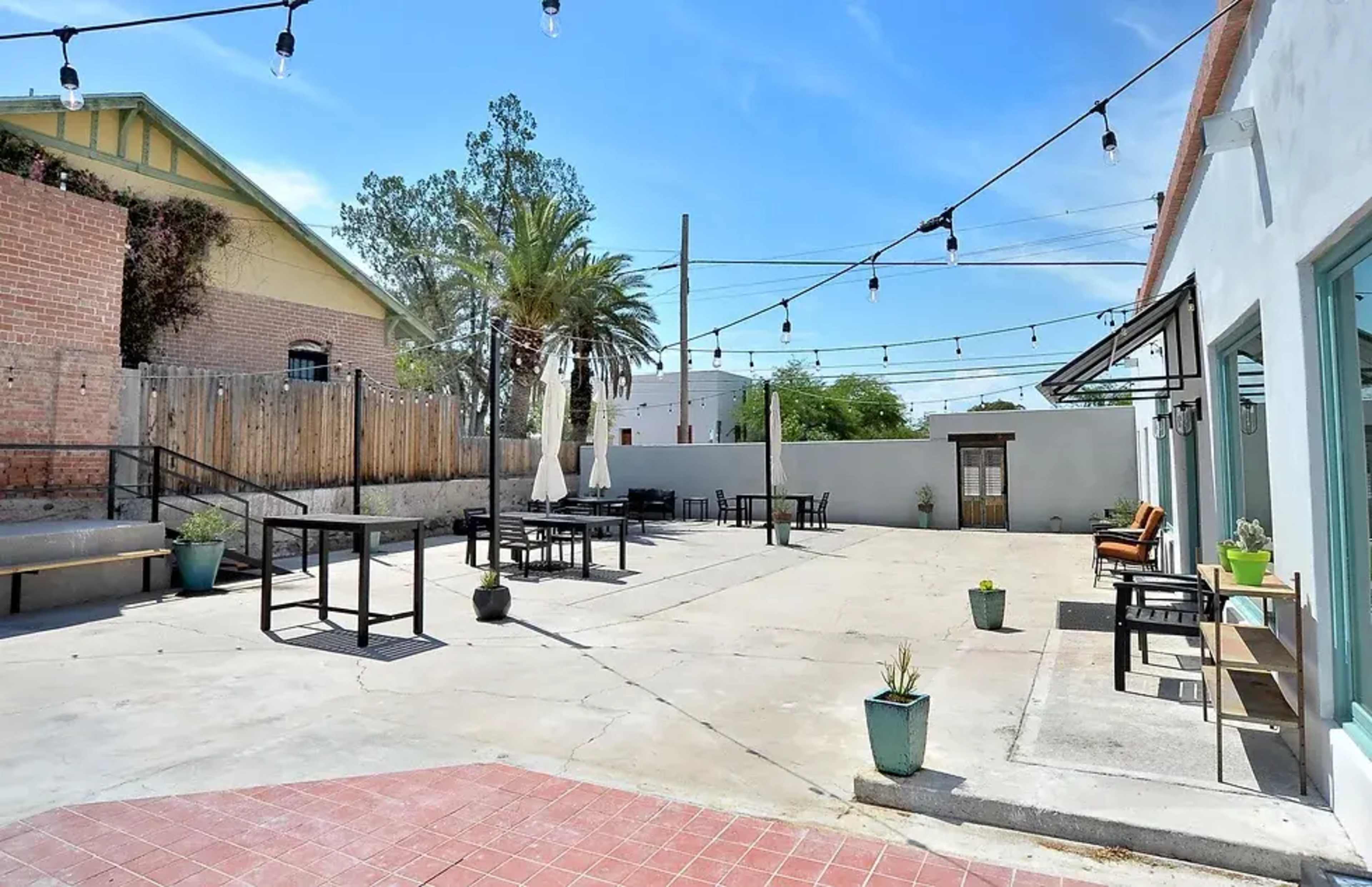 The image shows an outdoor space with tables and chairs, string lights overhead, and a few pots with plants.
