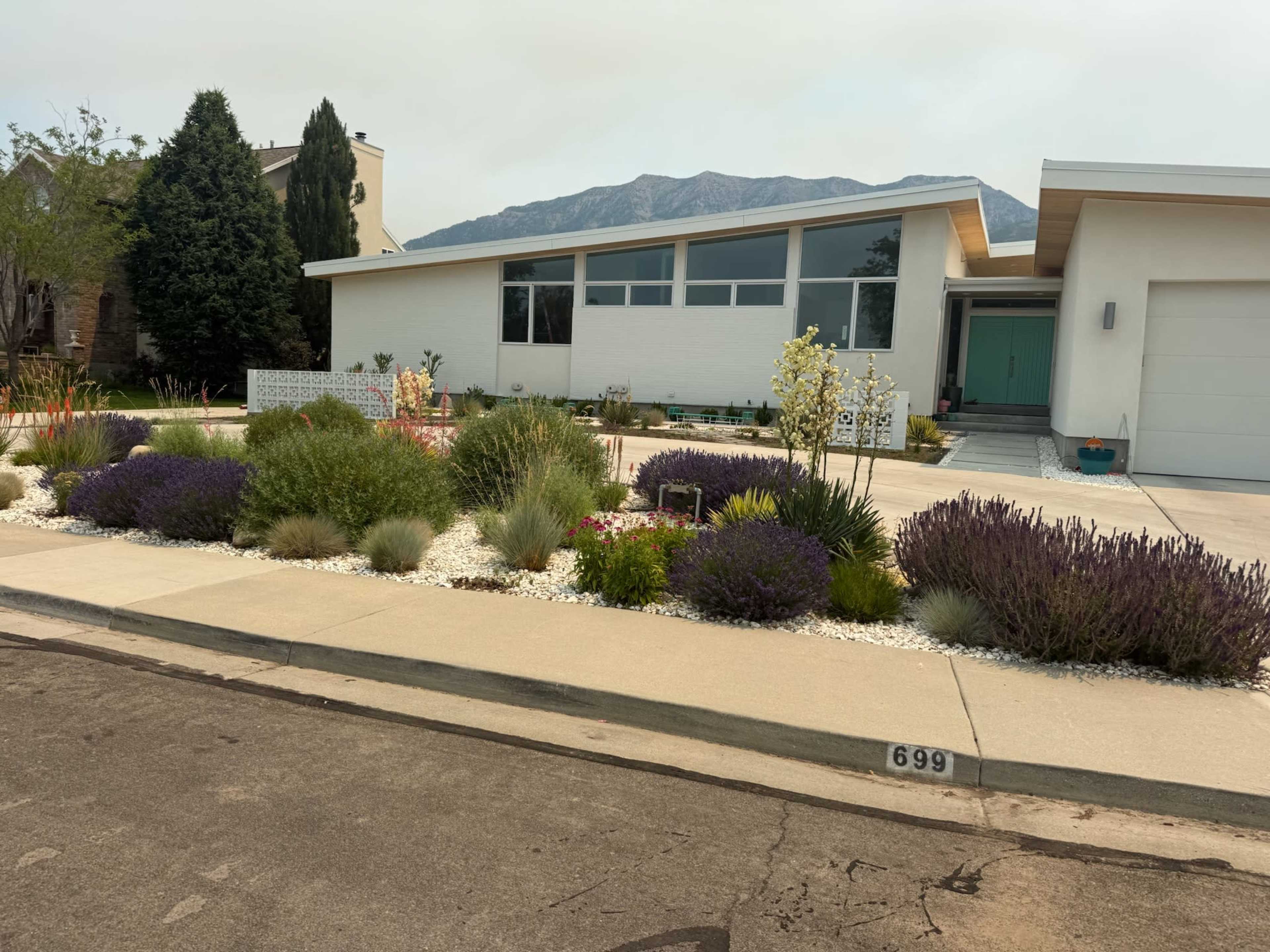 The image shows a modern house with a landscaped front yard featuring various plants and flowers, situated on a street with visible house numbers.