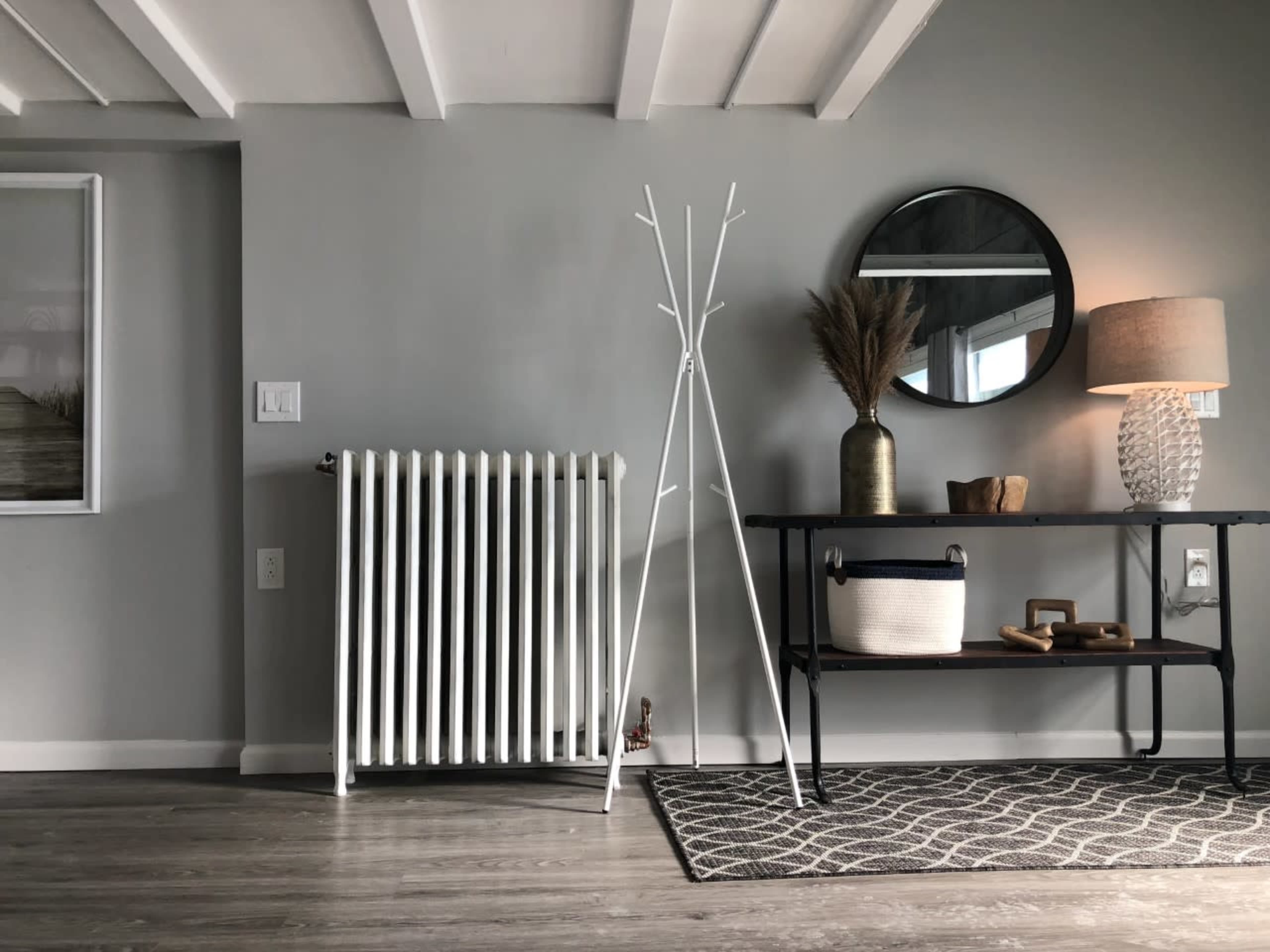 A minimalist interior with a white radiator, a coat stand, a round mirror, a lamp, and a table displaying a decorative basket and vase.