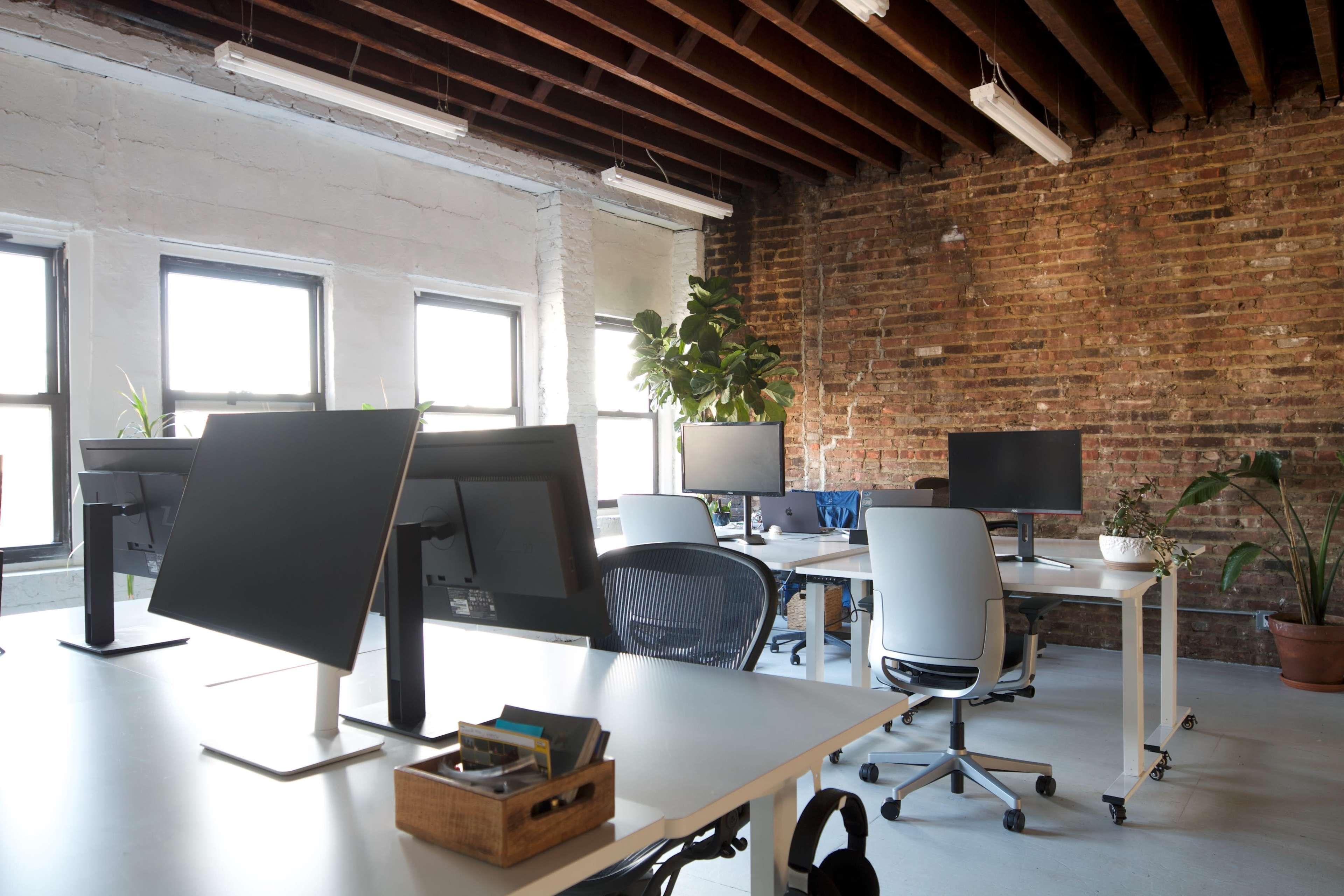 The image shows a modern office space featuring several desks with computer monitors, plants, and exposed brick walls.