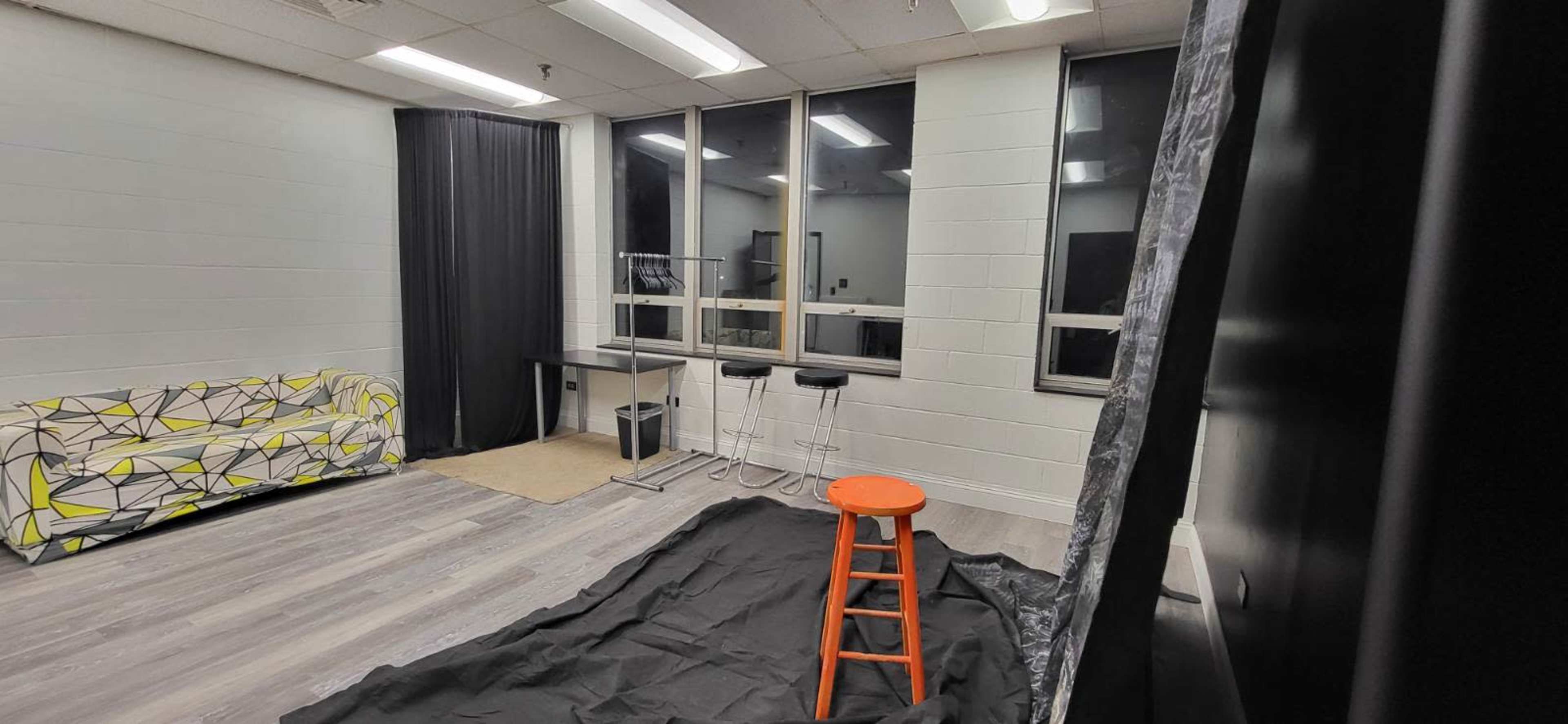 The image shows a room with a patterned couch, a tall orange stool, and a table against the wall, with a black draped fabric on the floor and large windows in the background.