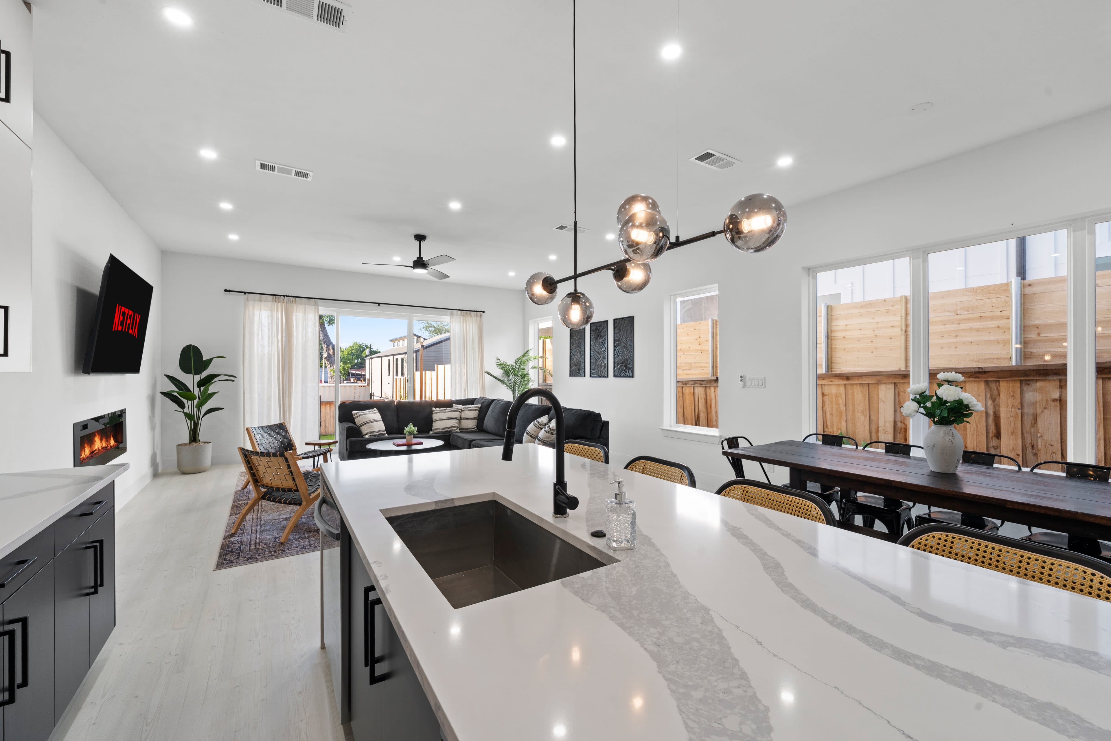 The image shows a modern open-concept living space that includes a kitchen with a large island, a dining area, and a lounge with a view of an outdoor patio.