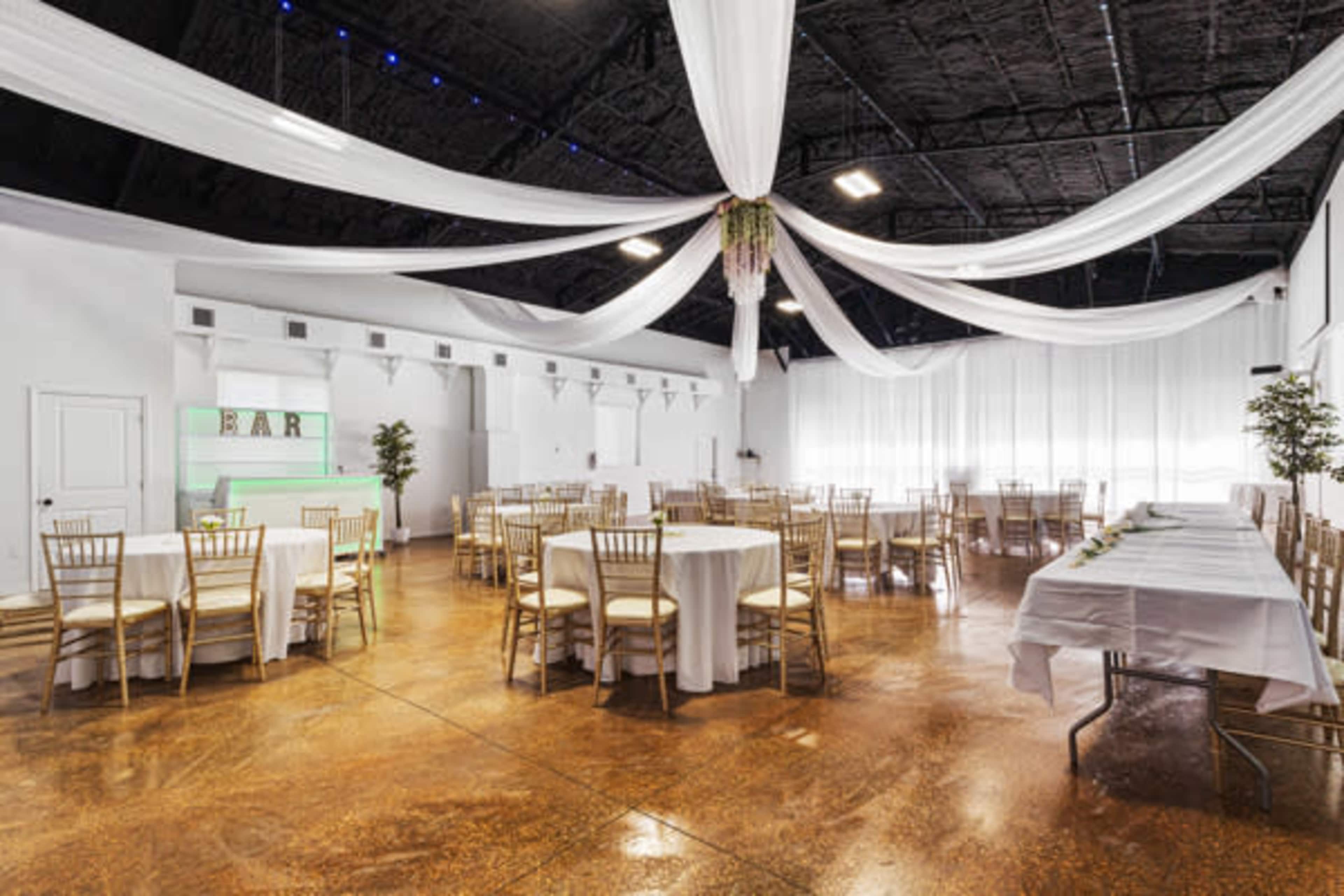 The image shows a spacious event venue with round tables and chairs arranged, draped ceilings, and a bar area illuminated in the background.