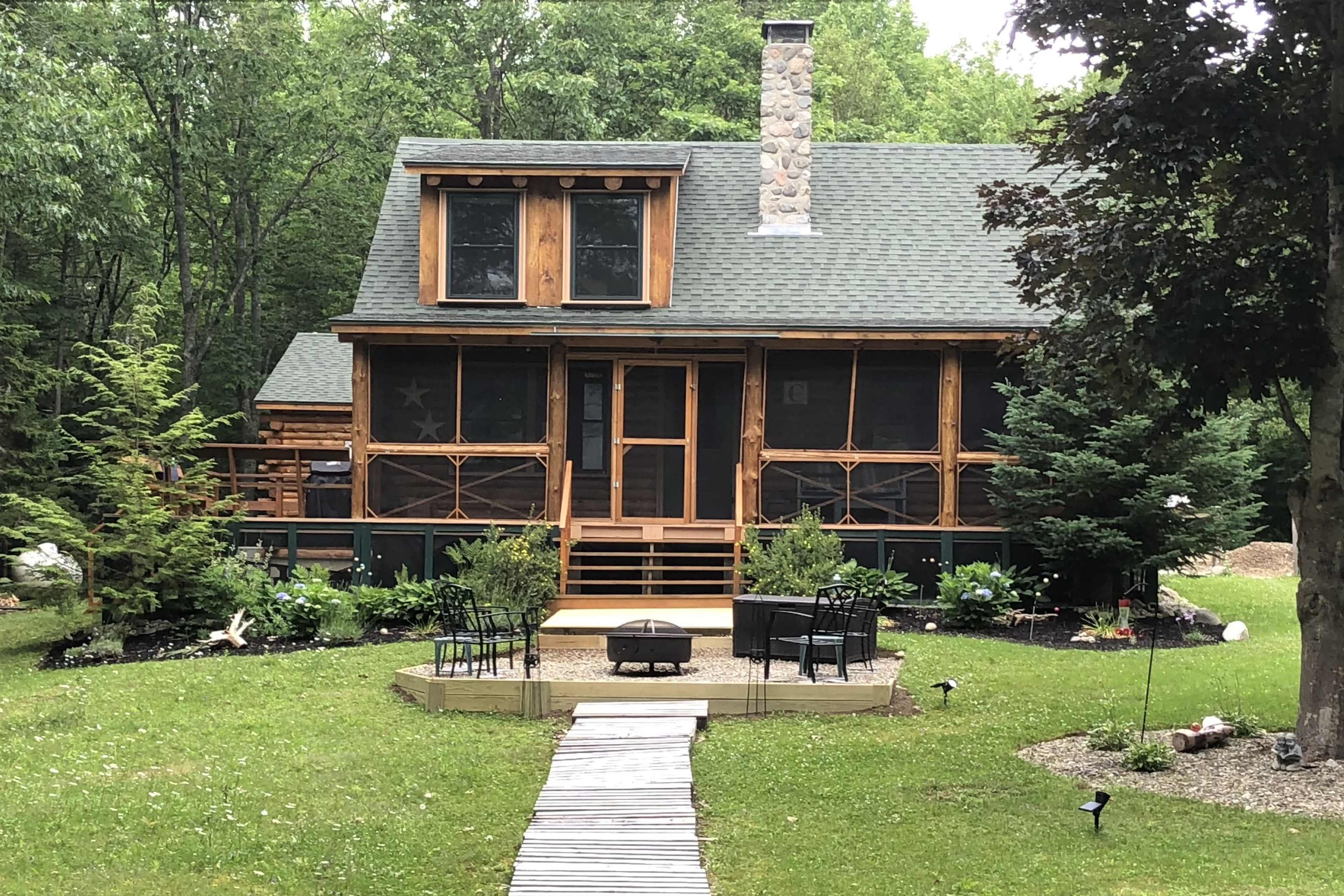 A rustic wooden cabin surrounded by greenery features a screened porch and a central outdoor fire pit with seating.