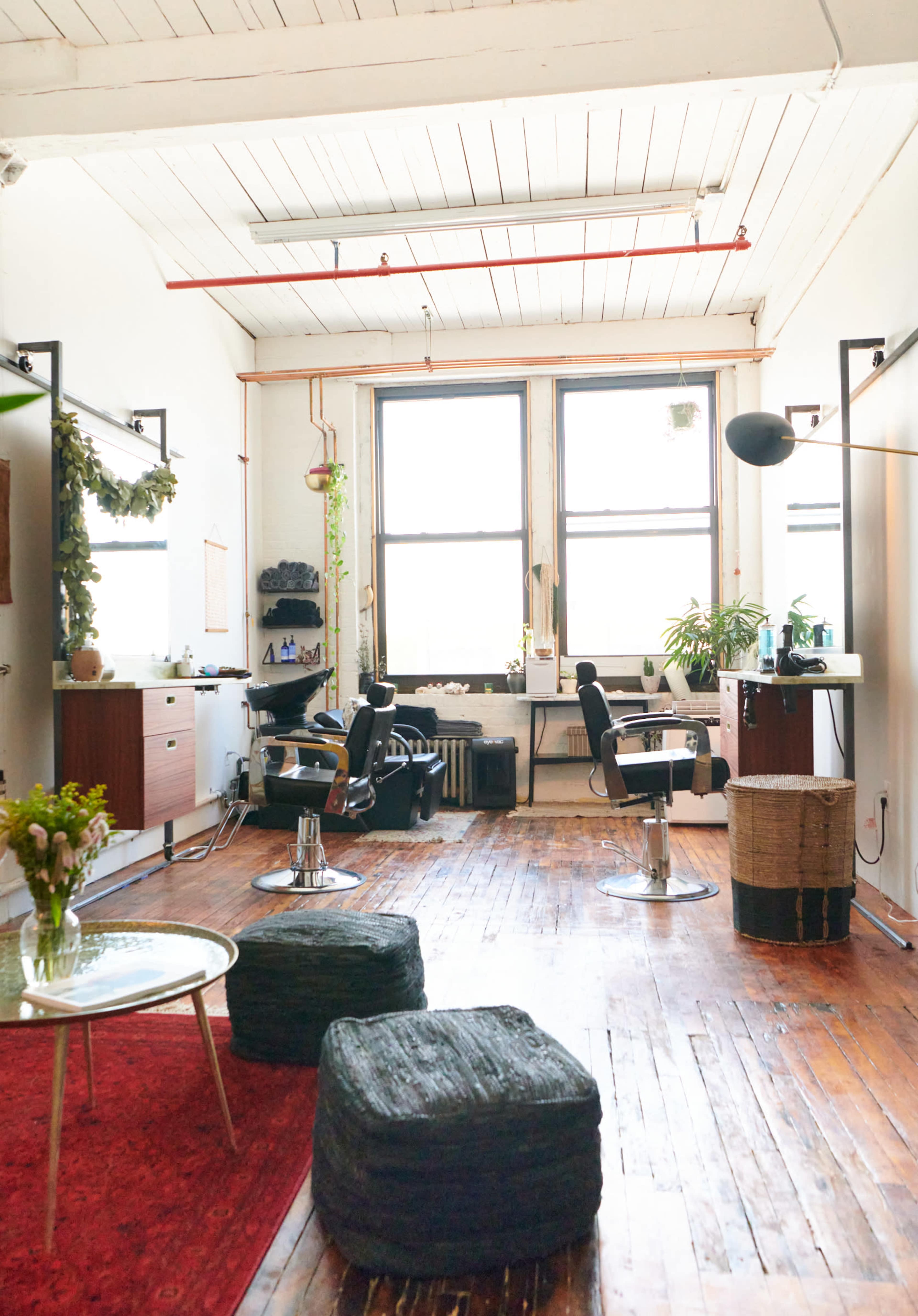 A bright, open salon space with wooden floors, large windows, and several styling stations equipped with black chairs.