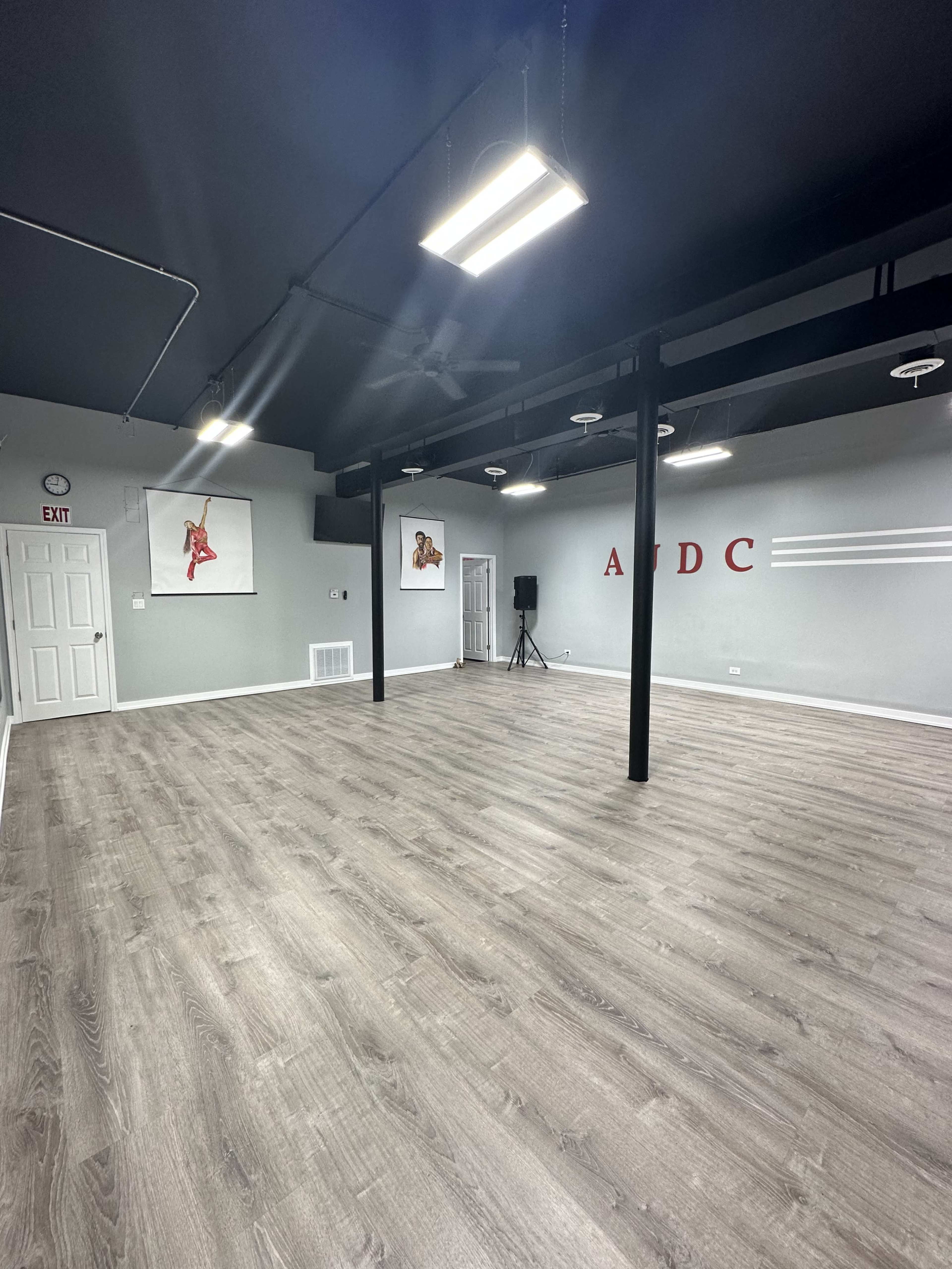The image shows a spacious dance studio with gray walls, wooden floors, and minimal decor, featuring an exit sign and artwork on the walls.