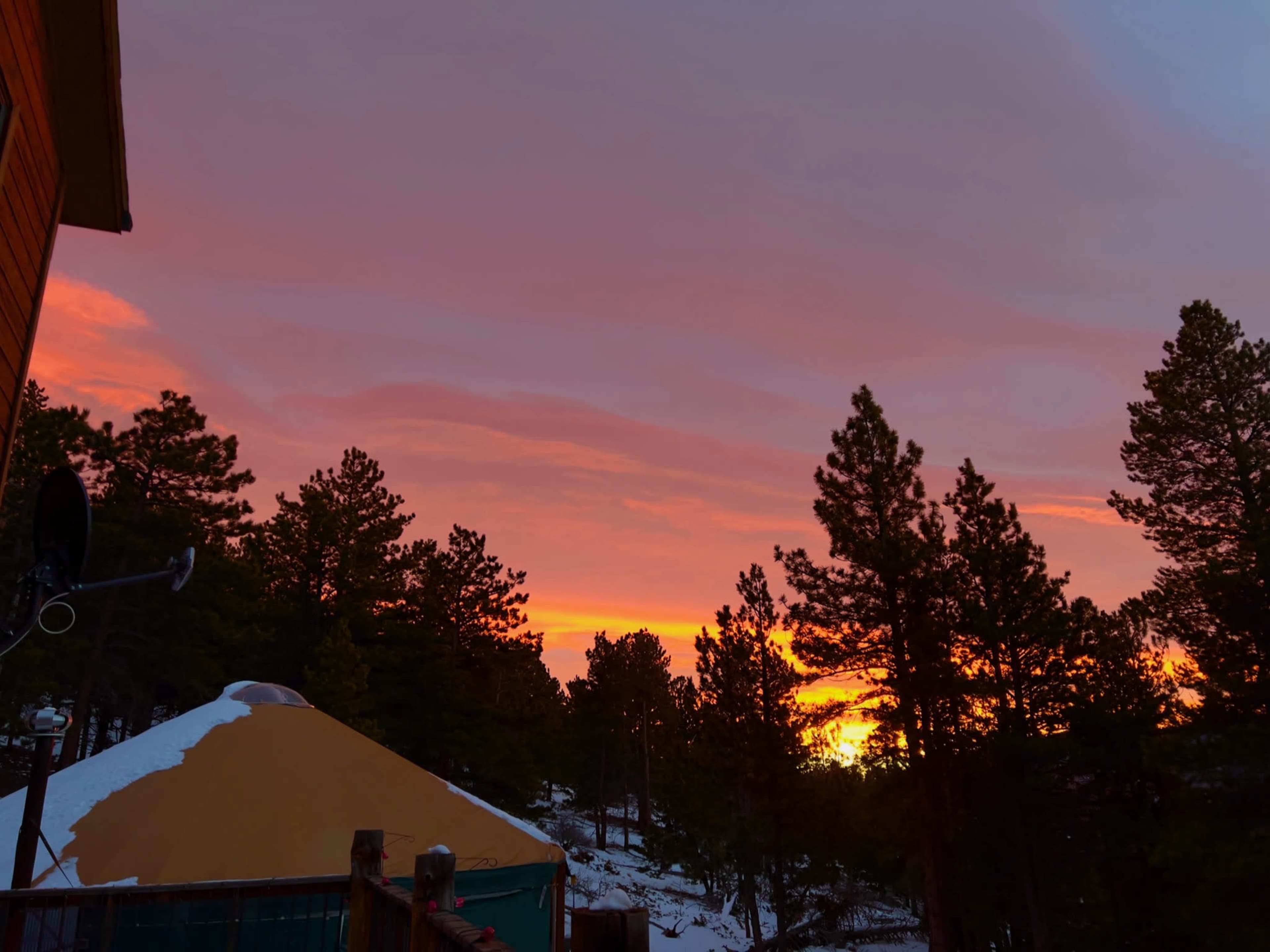 A colorful sunset illuminates the sky above a snowy landscape dotted with pine trees and a yurt.