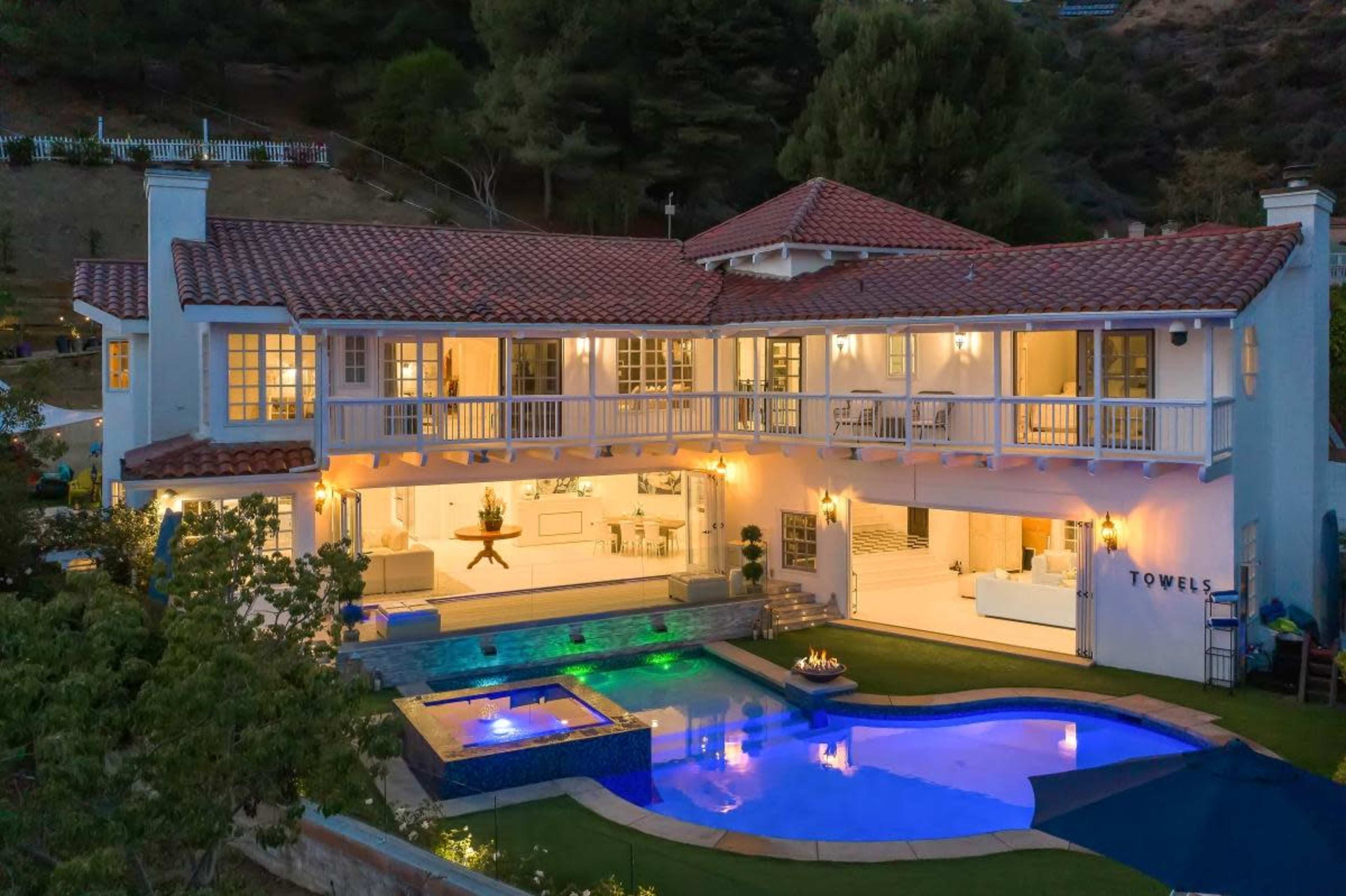 A two-story house with a tiled roof features a pool and outdoor seating area, illuminated in the evening.