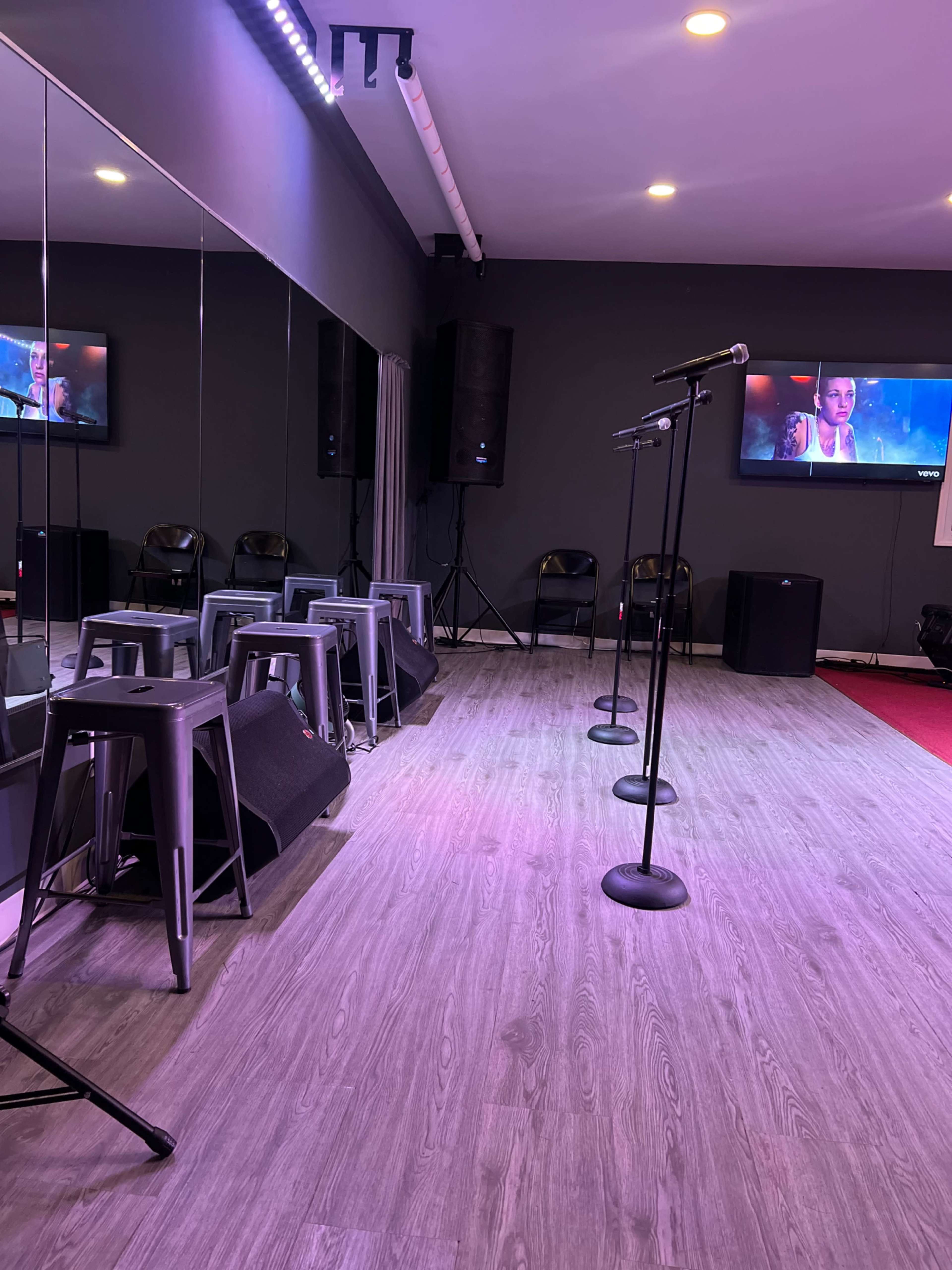 A small performance space features several microphones on stands, stools, and monitors displaying video content.