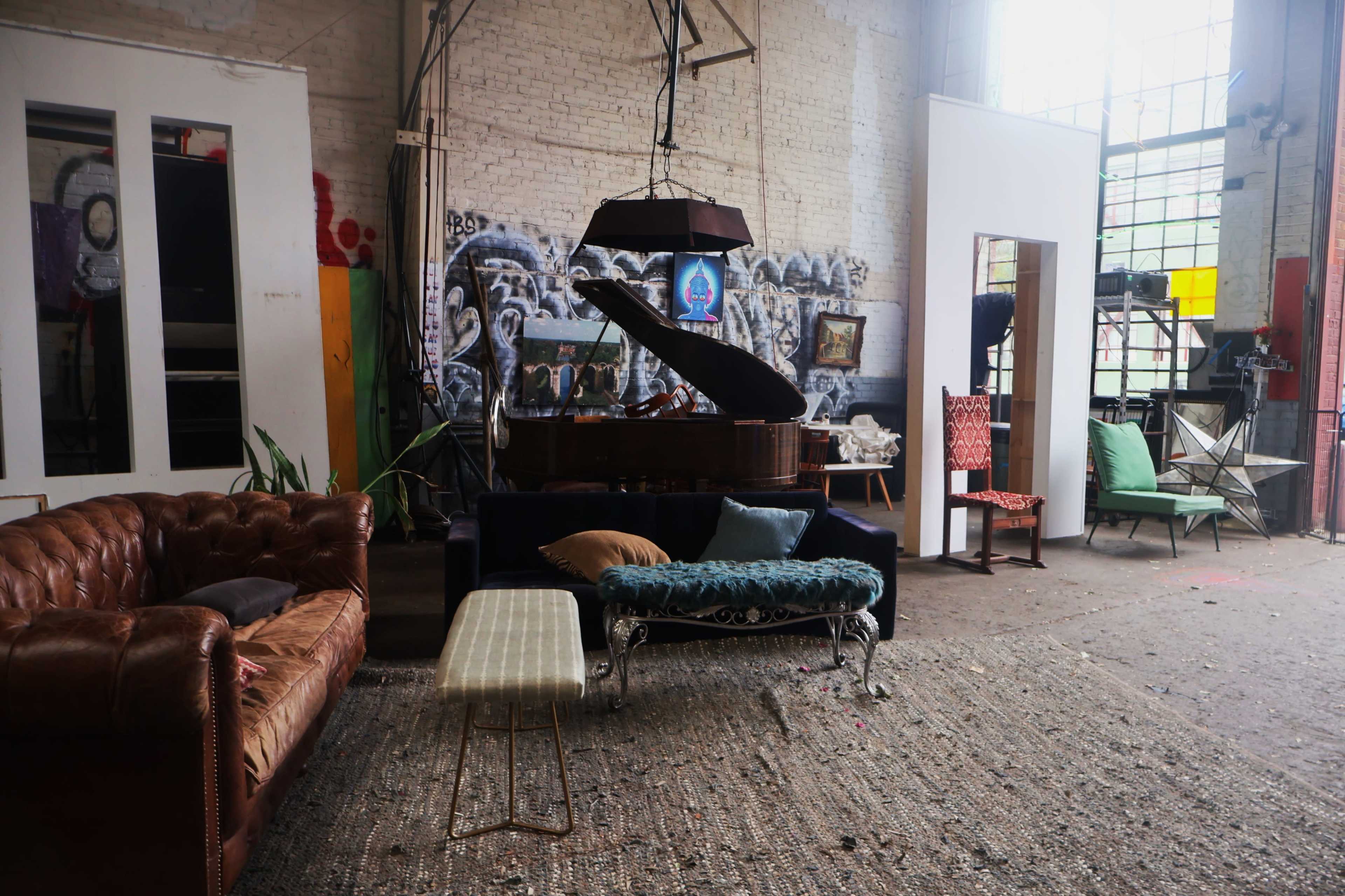 The image shows an eclectic interior of an industrial space featuring a piano, various seating arrangements, a large mirror, and artwork on the walls.