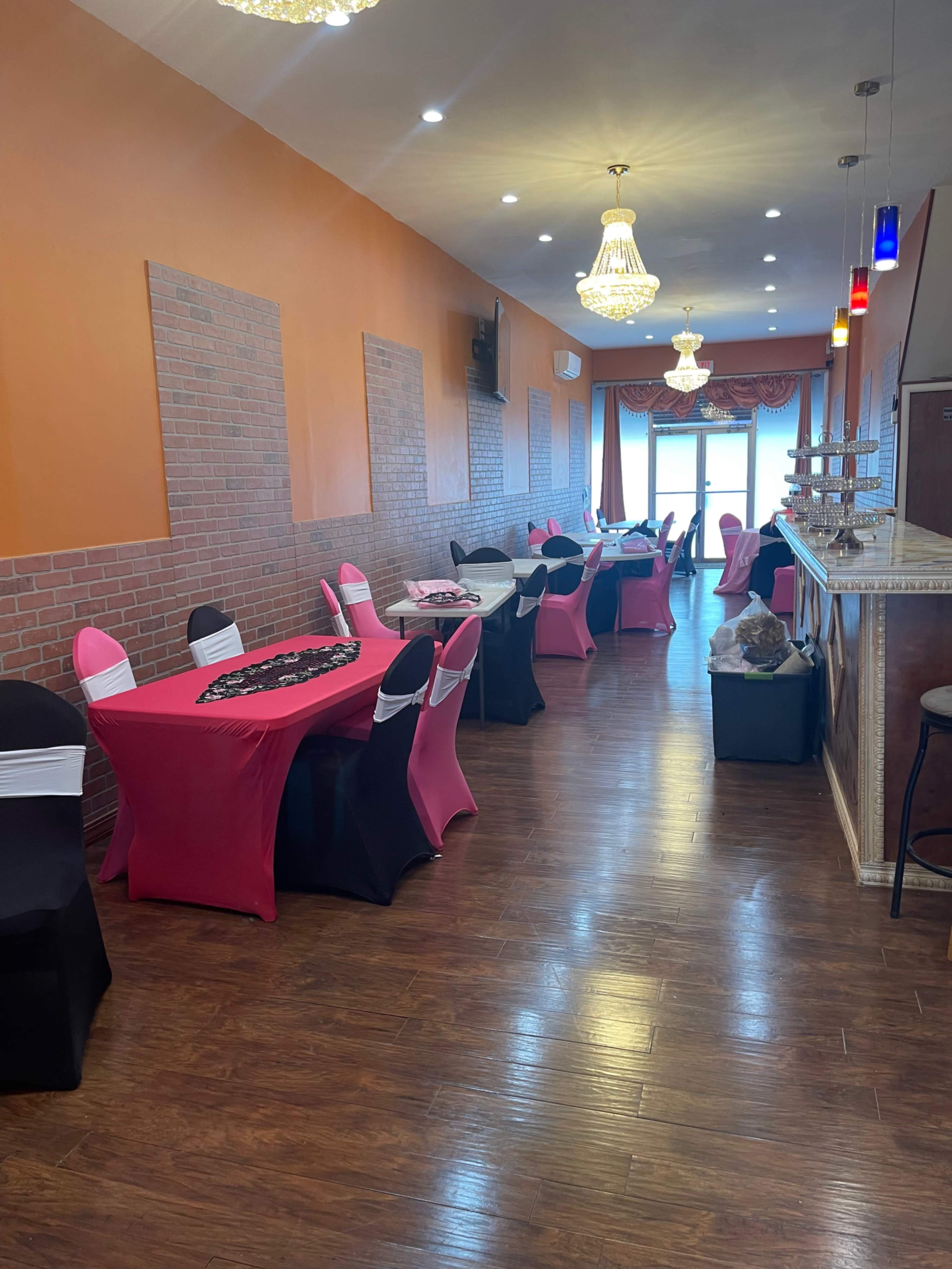 The interior of a restaurant features rows of tables dressed in black and pink tablecloths, with decorative lighting and a bar at the far end.