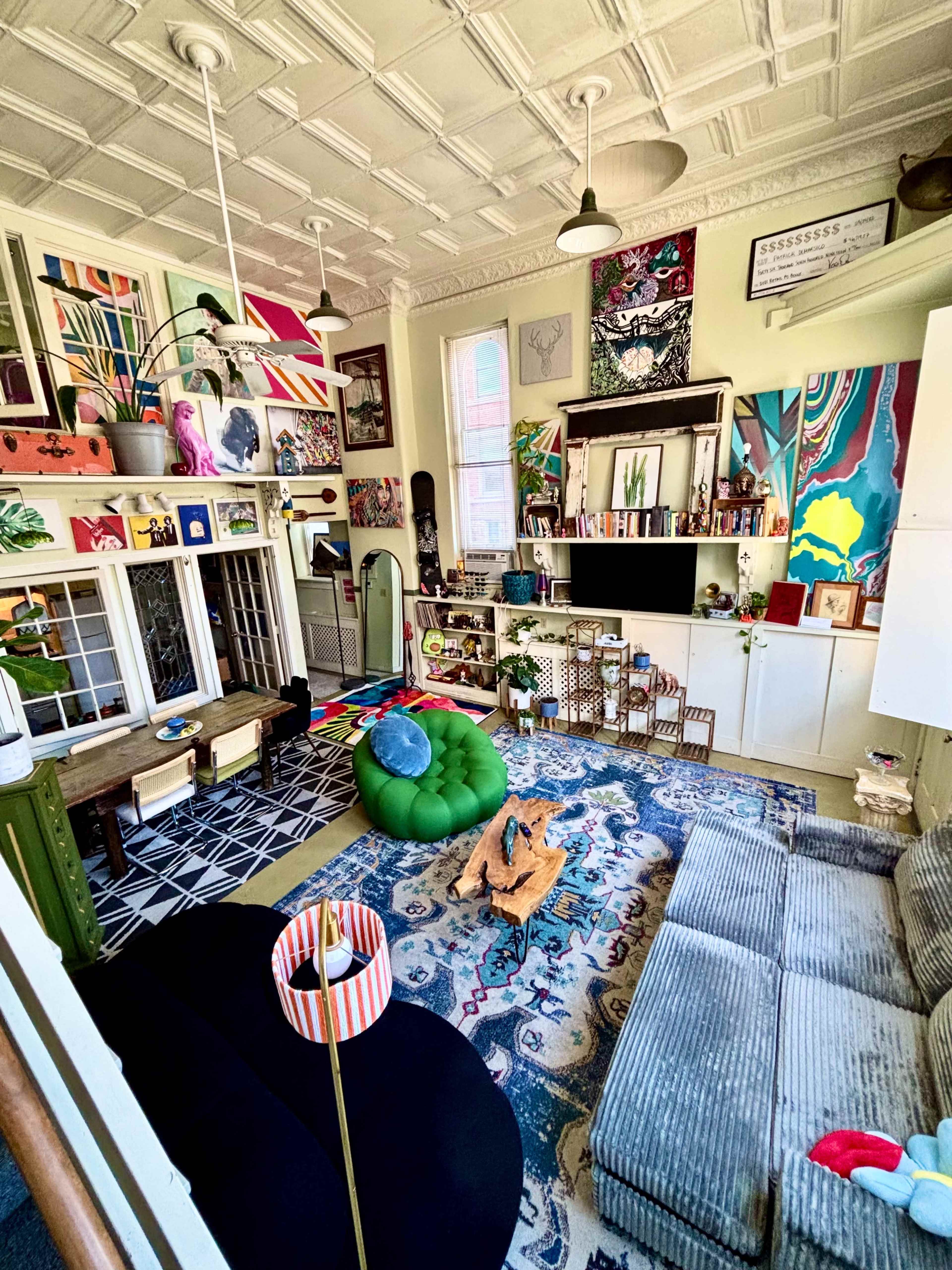 The room features a mix of vibrant artwork on the walls, colorful furniture including a green bean bag and a gray sofa, and a variety of decorative items arranged throughout the space.