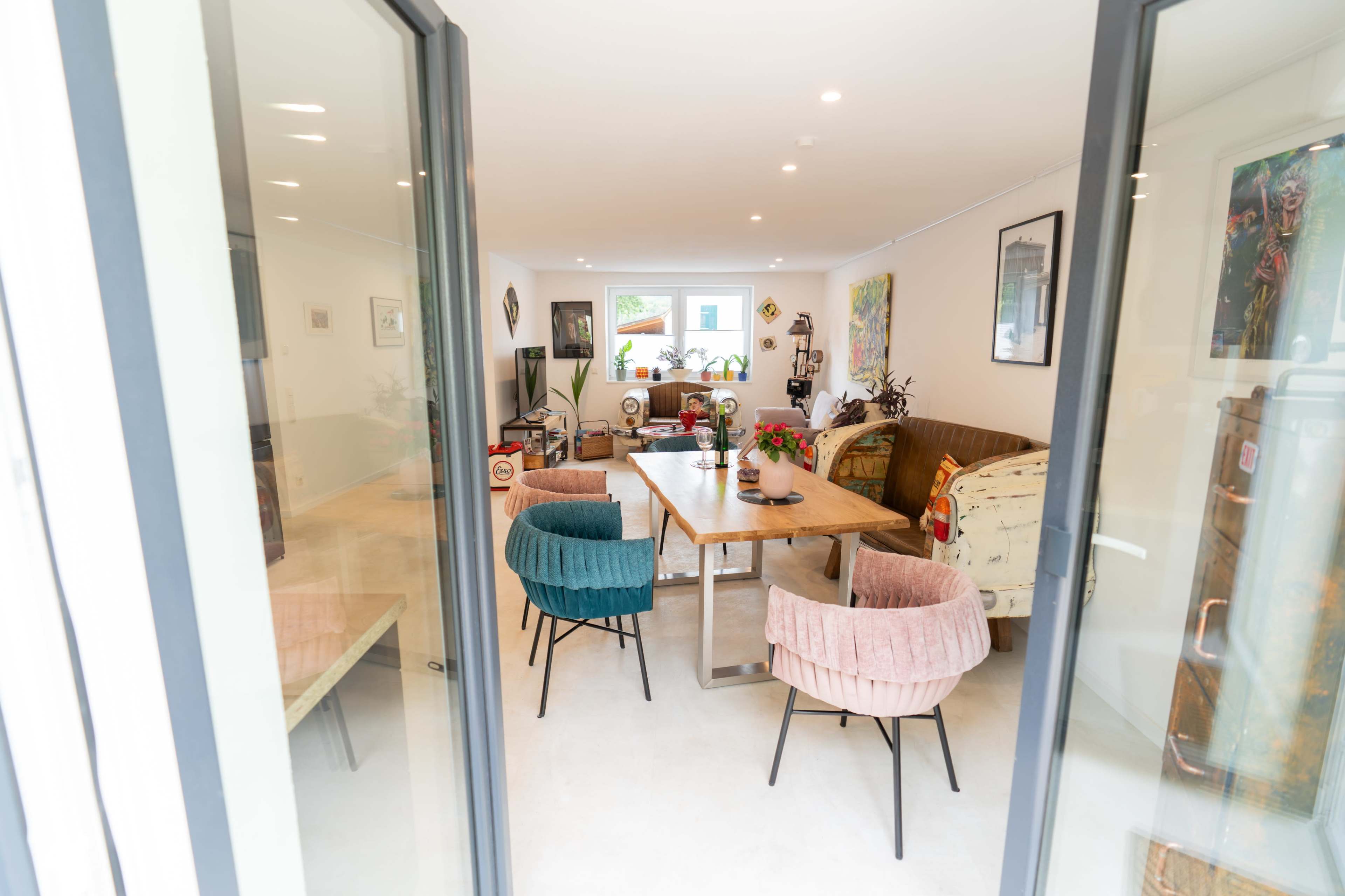 The image shows a modern living space with a dining table surrounded by colorful chairs, artwork on the walls, and various decorative elements.