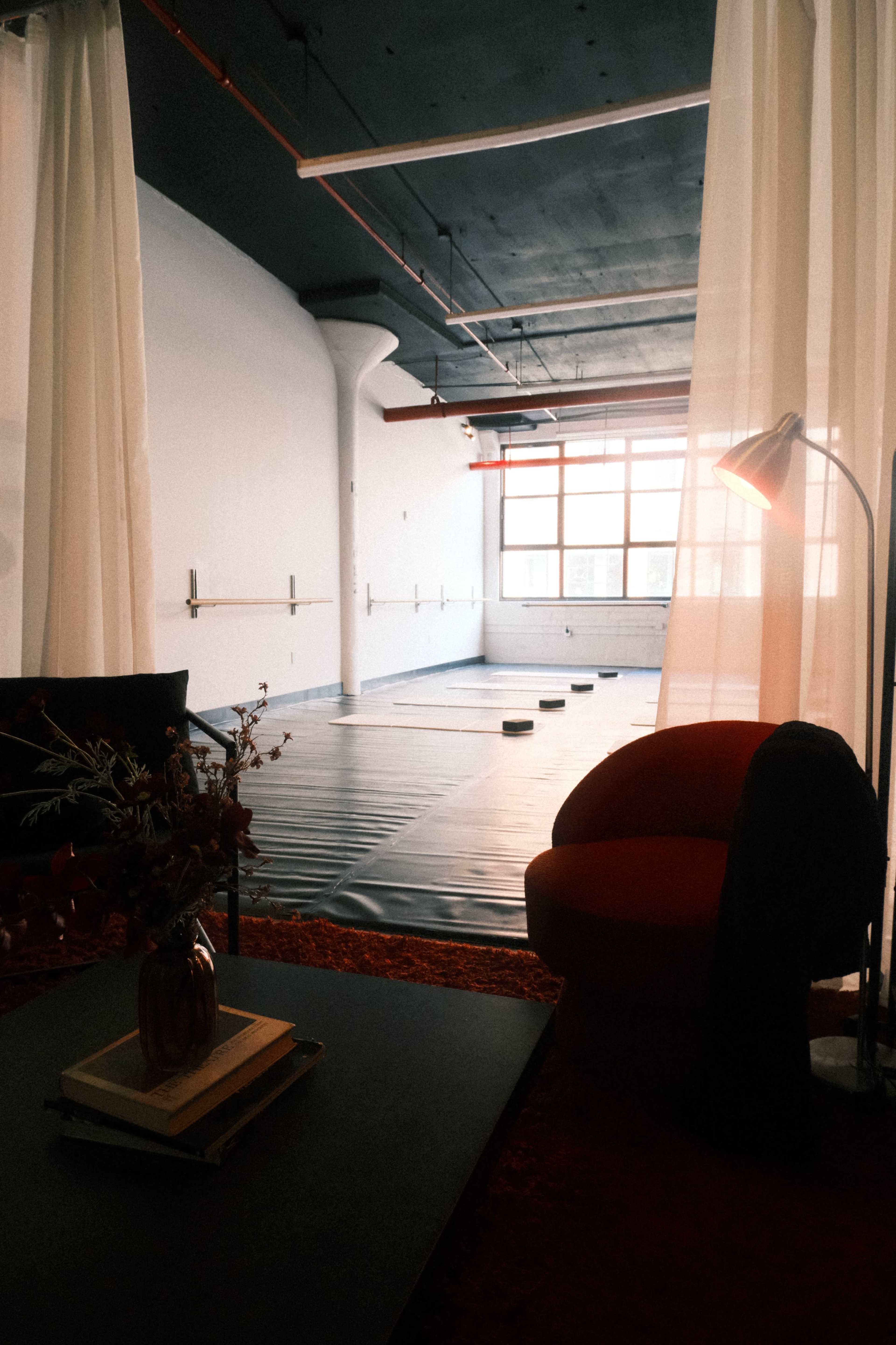 The image shows a spacious, minimalist studio with large windows, a black floor mat, and a red chair next to a small table with a vase and books.