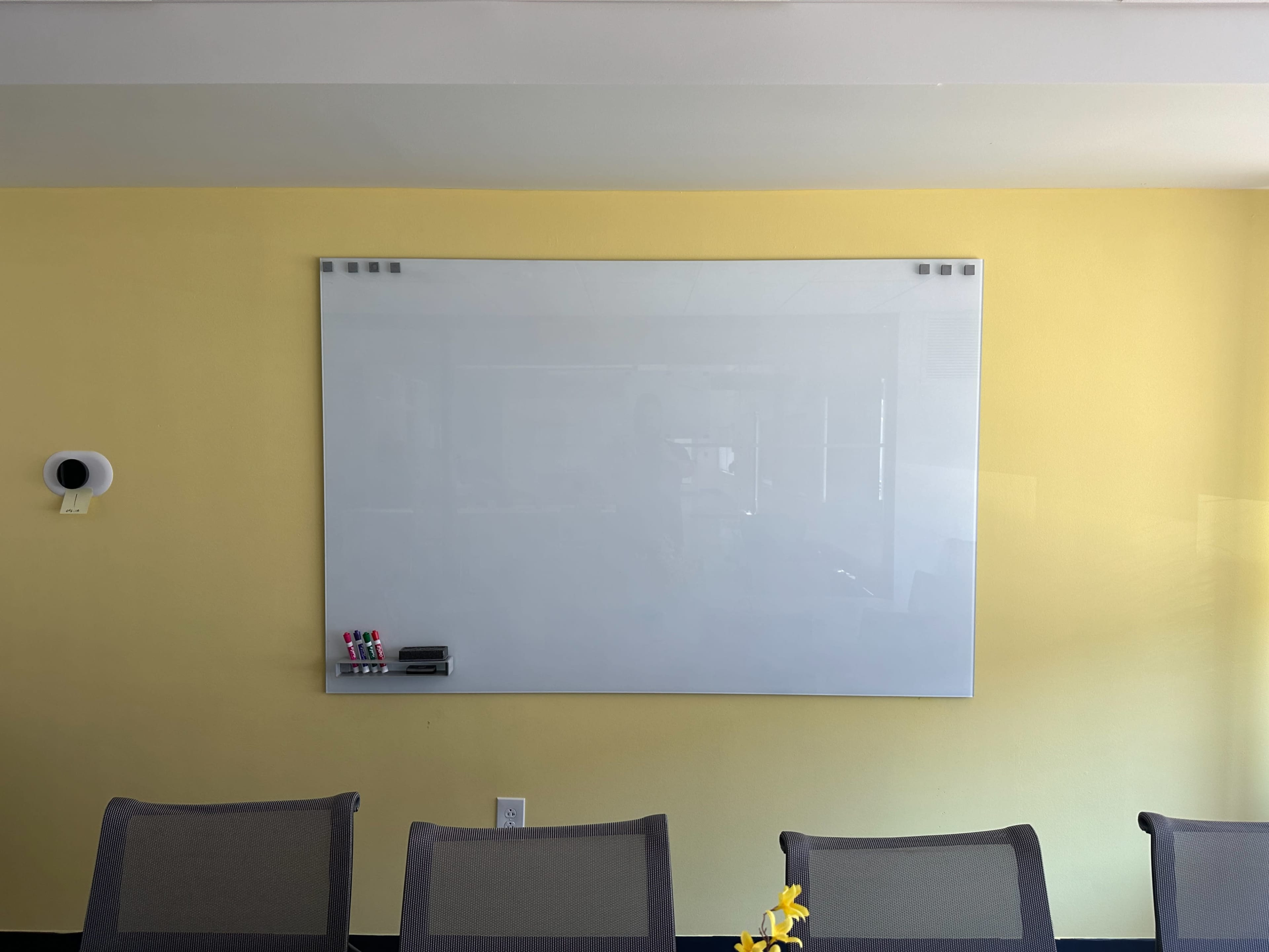 A blank whiteboard mounted on a yellow wall, with a row of empty chairs in front.