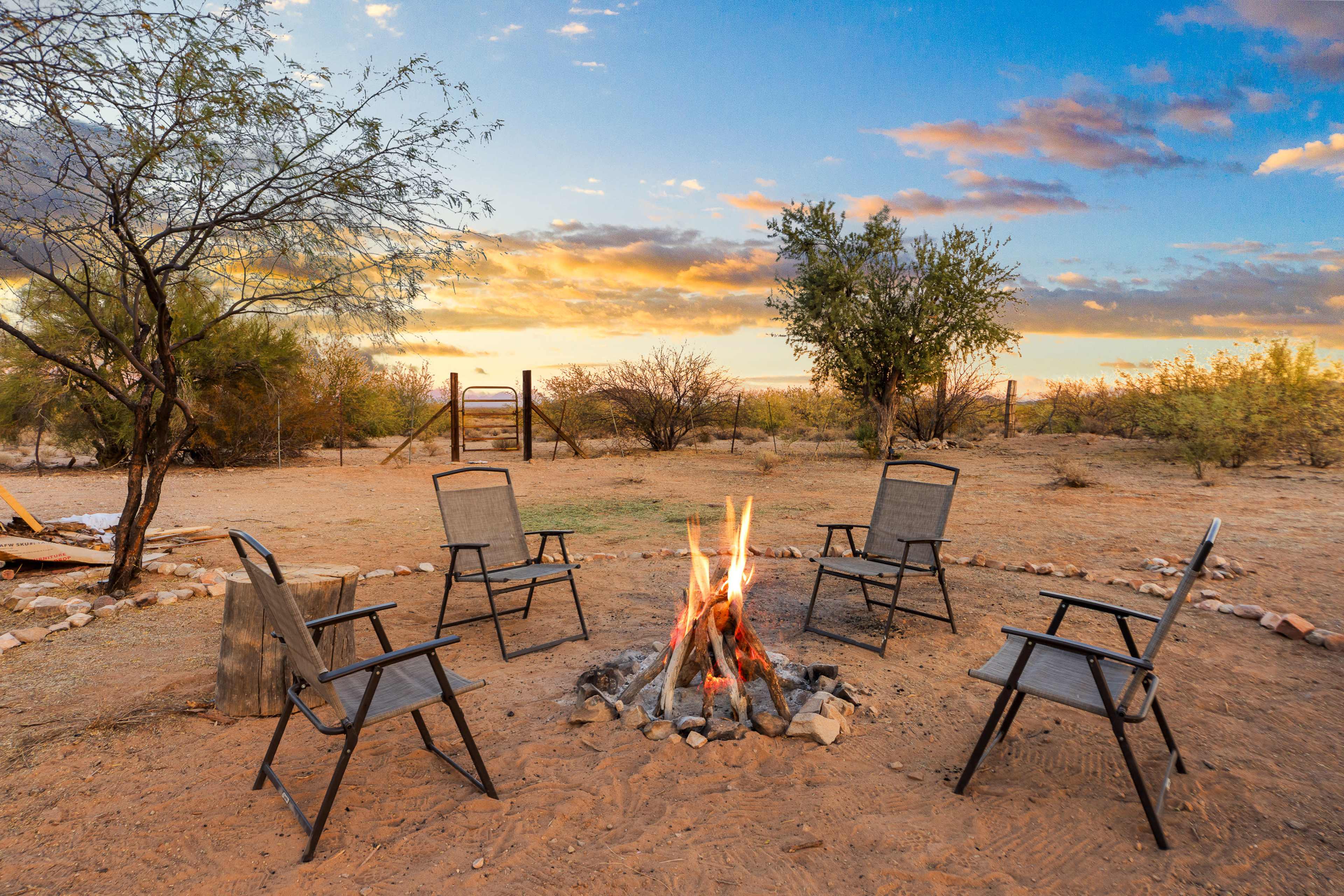 A campfire surrounded by five chairs is set in a desert landscape under a colorful sunset.