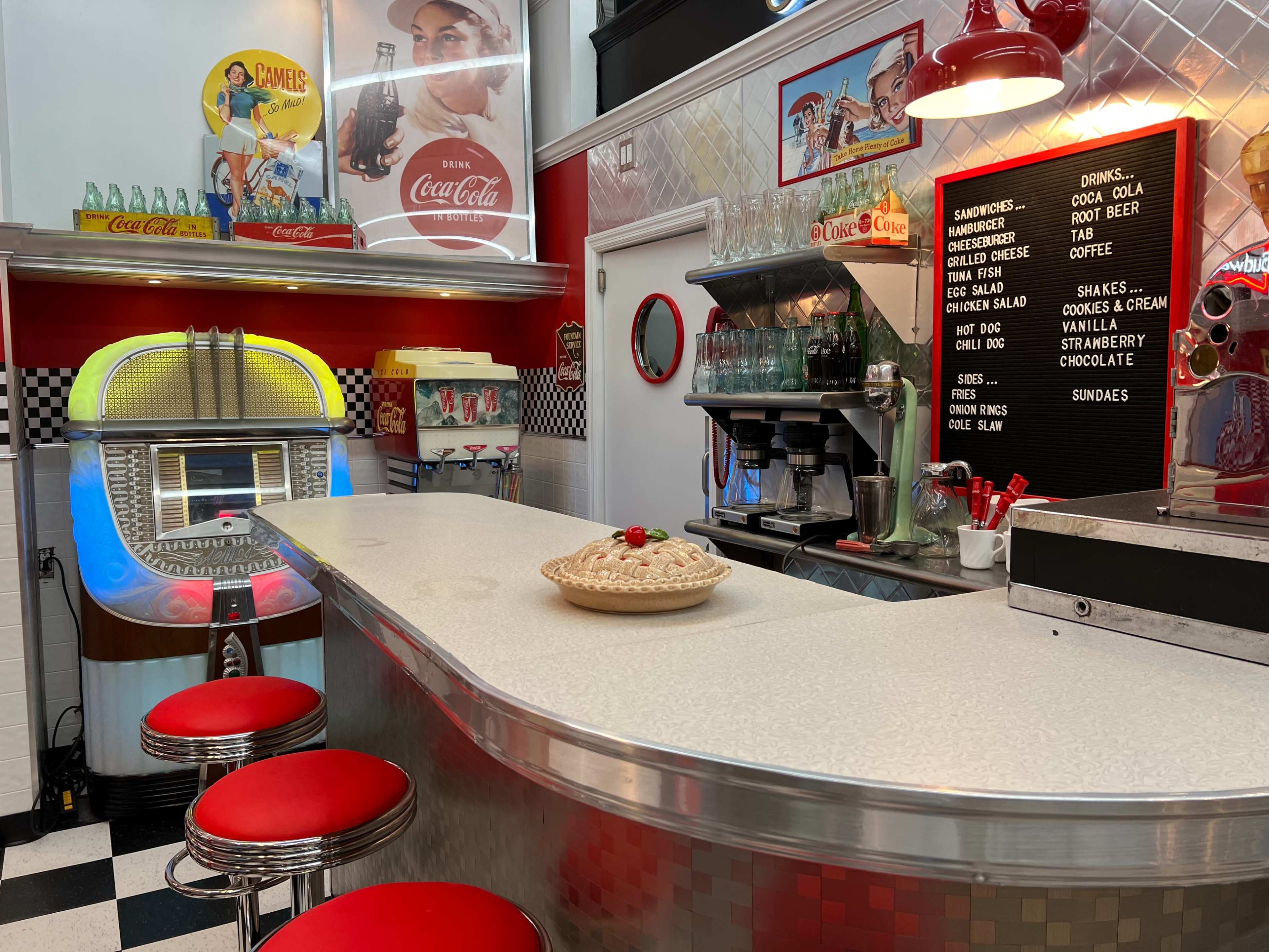 A retro diner interior with a rounded counter, red bar stools, a jukebox, and a menu on the wall featuring desserts and drinks.