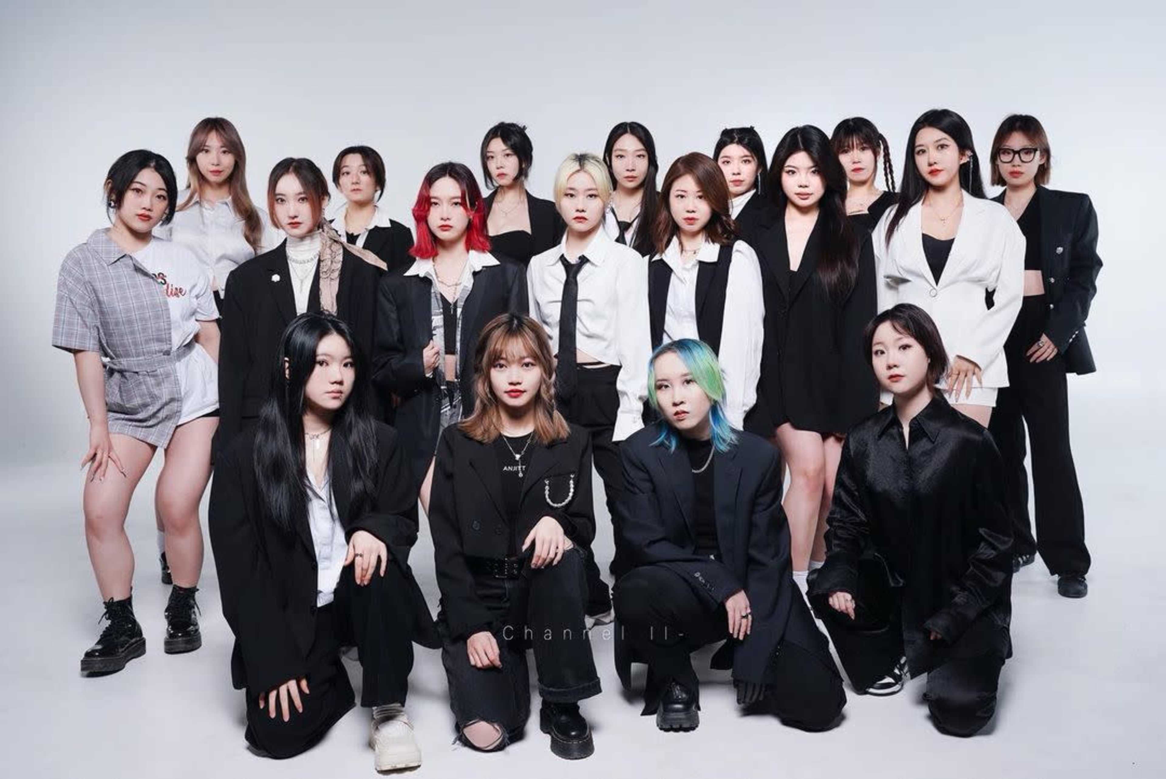 A group of 20 individuals, dressed in various styles of formal and casual attire, poses together in a studio setting with a light background.