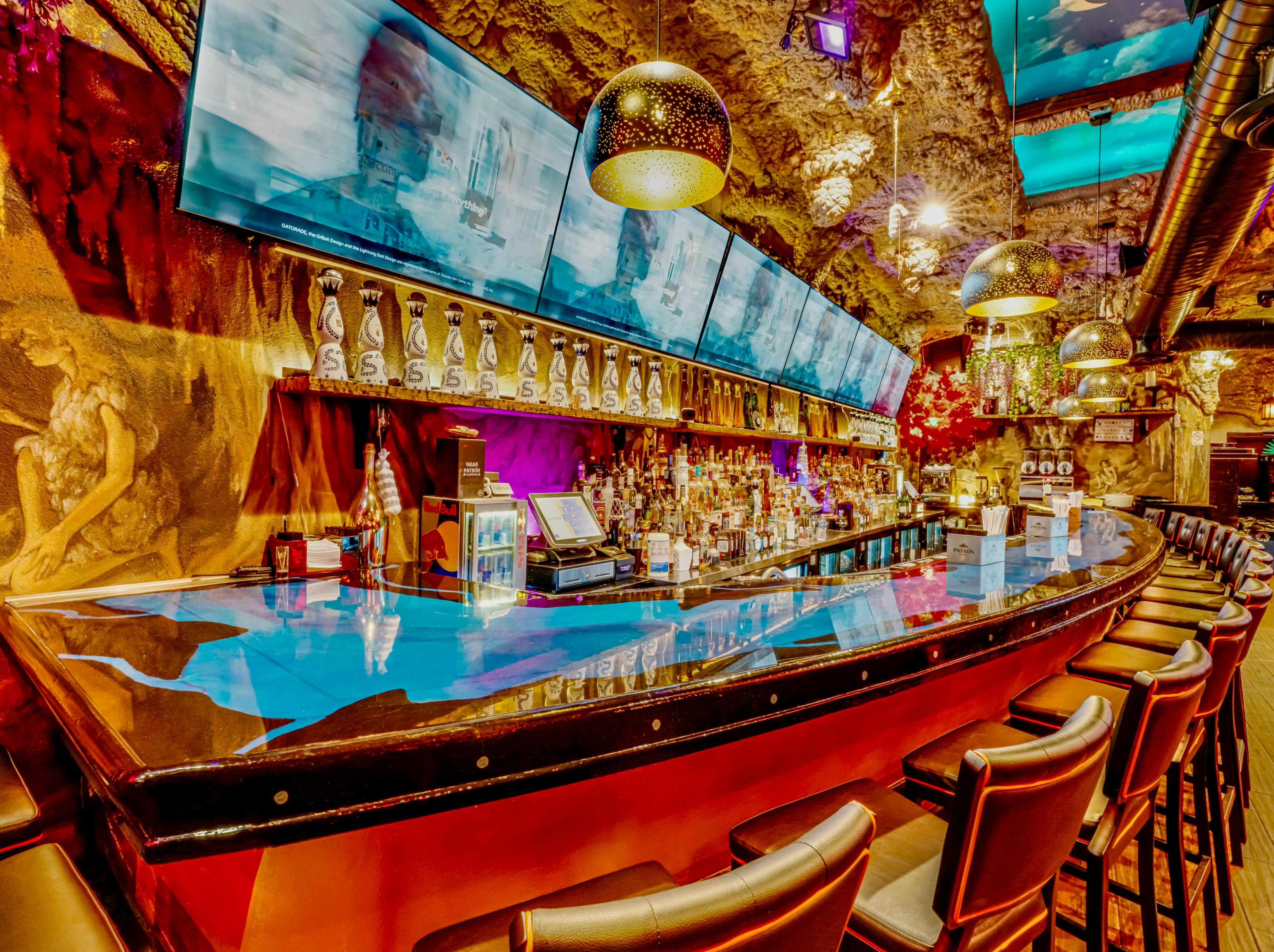 The image showcases a bar with a curved countertop, multiple large screens displaying visuals, and a variety of bar stools lined up along the counter in a dimly lit cave-like setting.