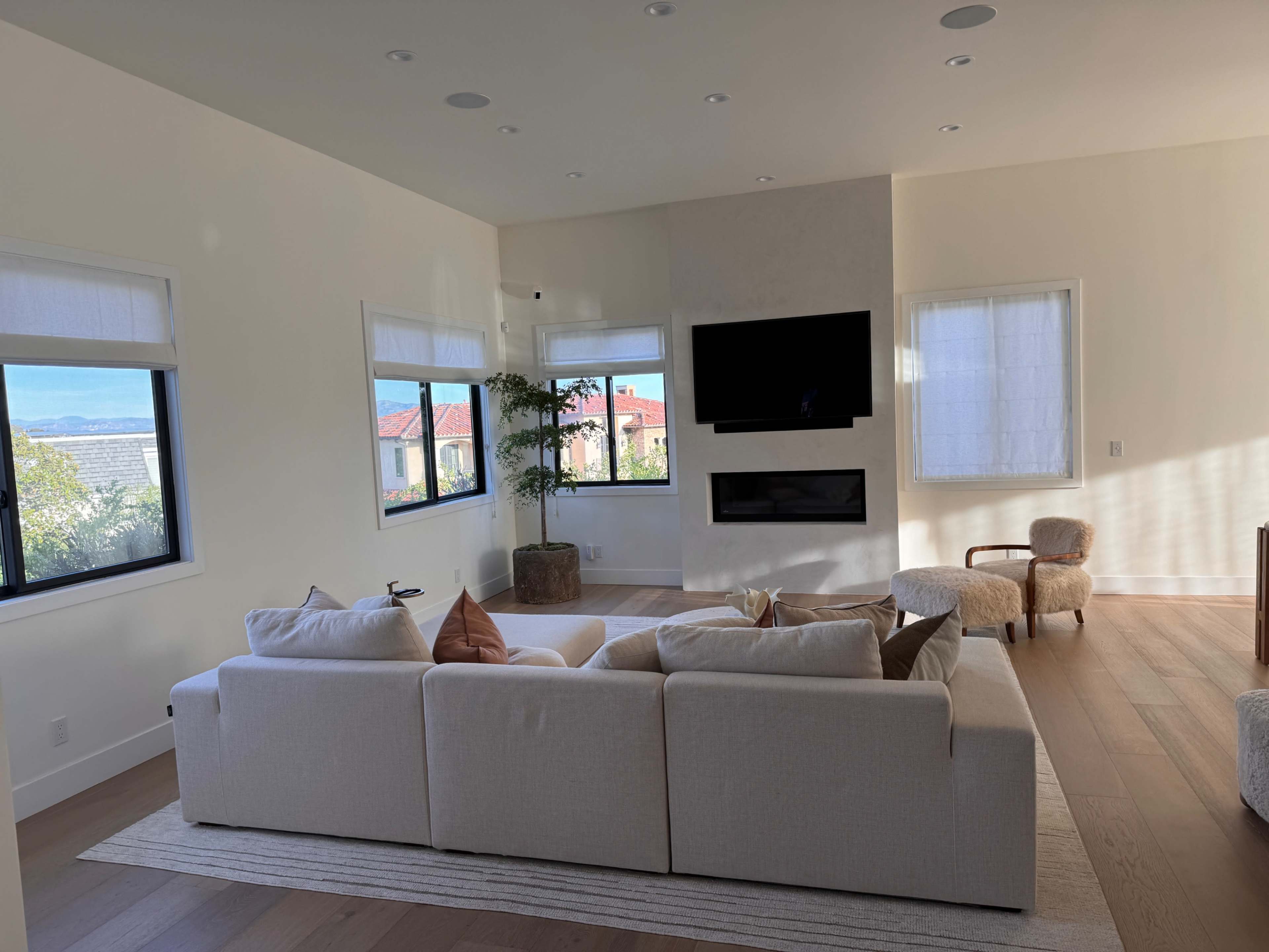 A light-colored living room with a large sectional sofa, a mounted TV on the wall, and a cozy armchair near a window.
