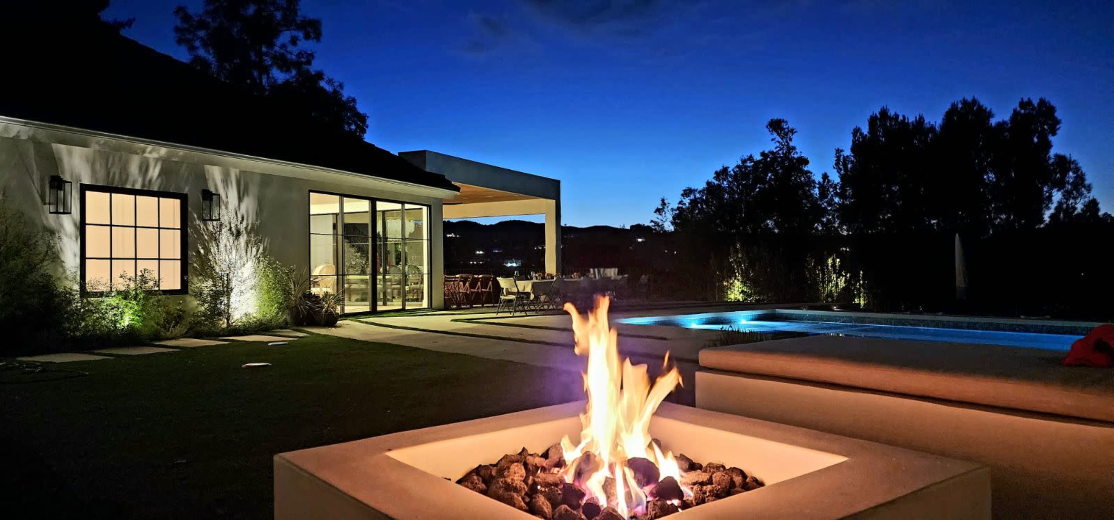 A fire pit with flames is situated in a backyard near a swimming pool, illuminated by the evening sky and surrounded by landscaped greenery.