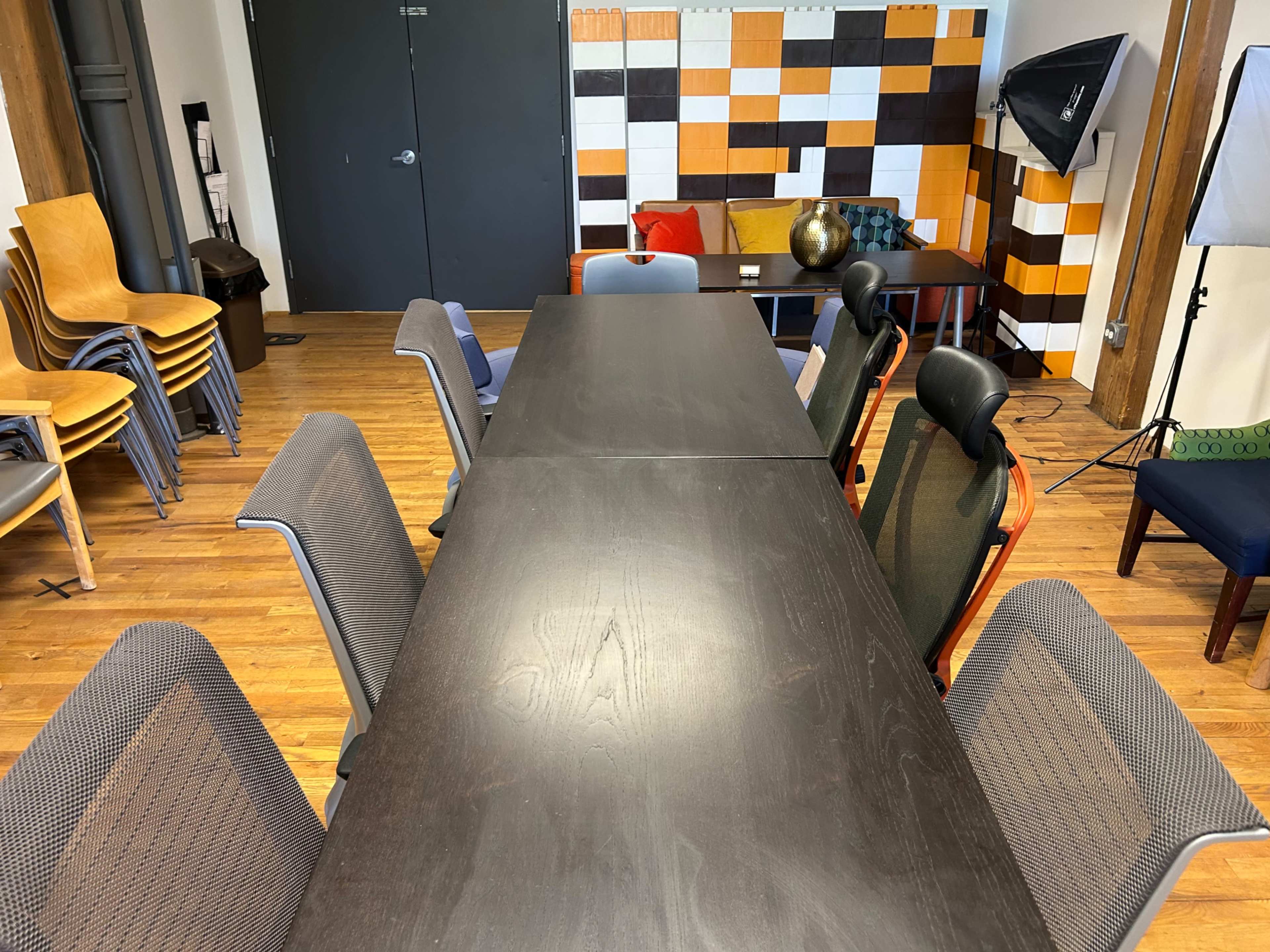 A long, dark wooden table surrounded by various office chairs sits in a room with a colorful tiled wall and stacked chairs in the background.