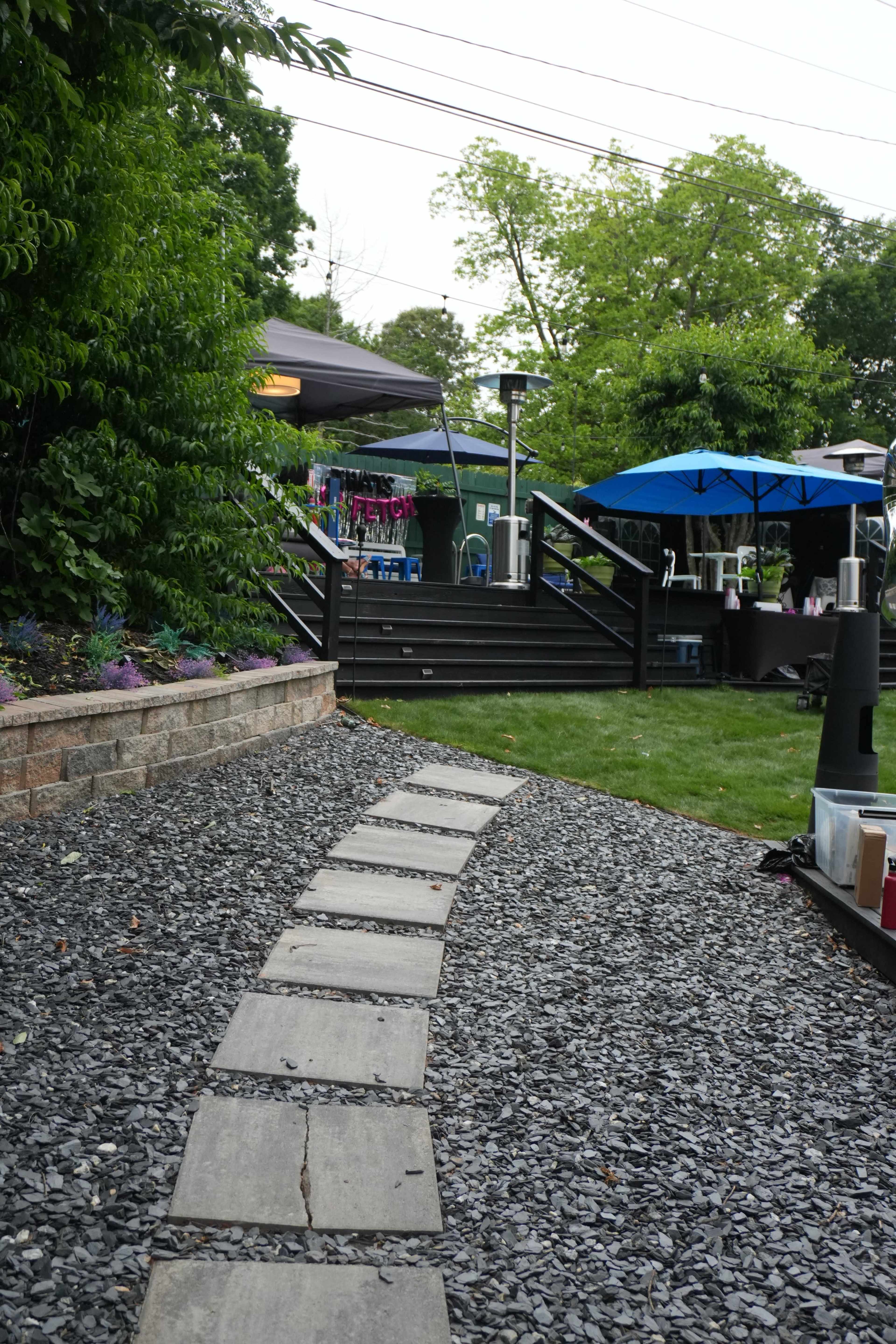 A gravel pathway lined with stone slabs leads toward a deck with umbrellas and tents in a backyard setting.