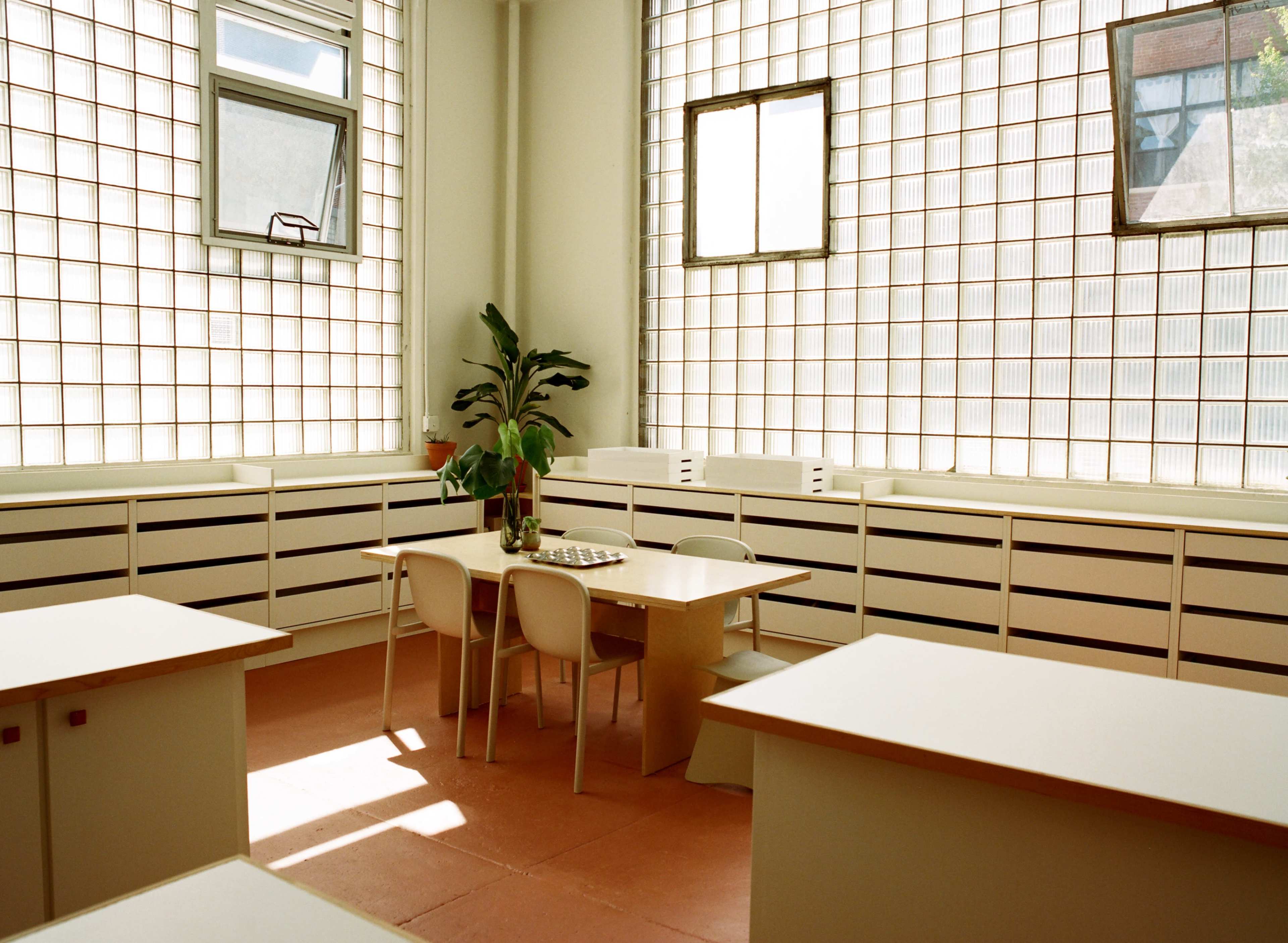 The image shows a bright, modern classroom featuring a table with chairs and large windows made of glass blocks that let in natural light.