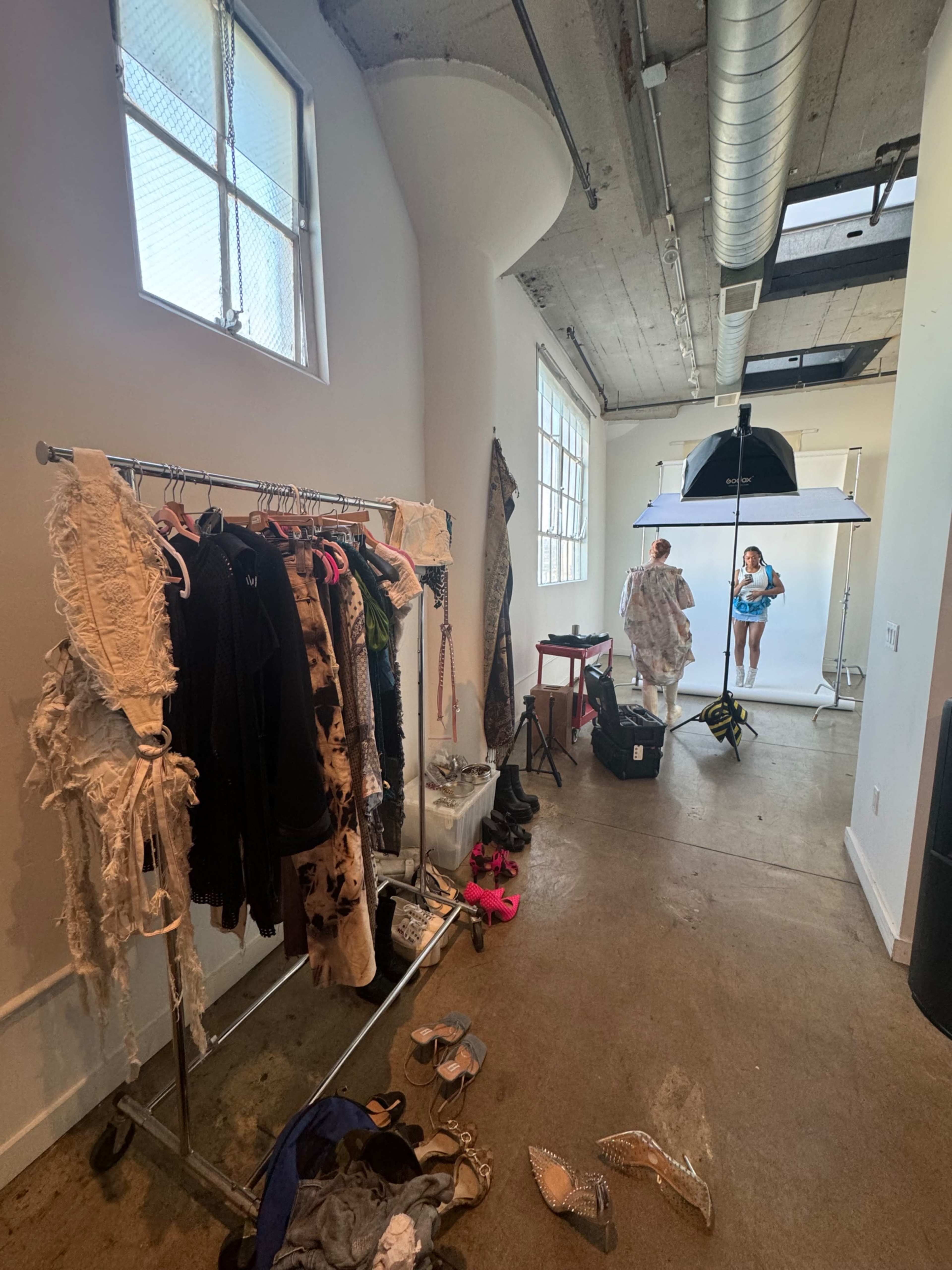 A photography studio with clothing racks on one side, a model posing in front of a backdrop, and various items scattered on the floor.