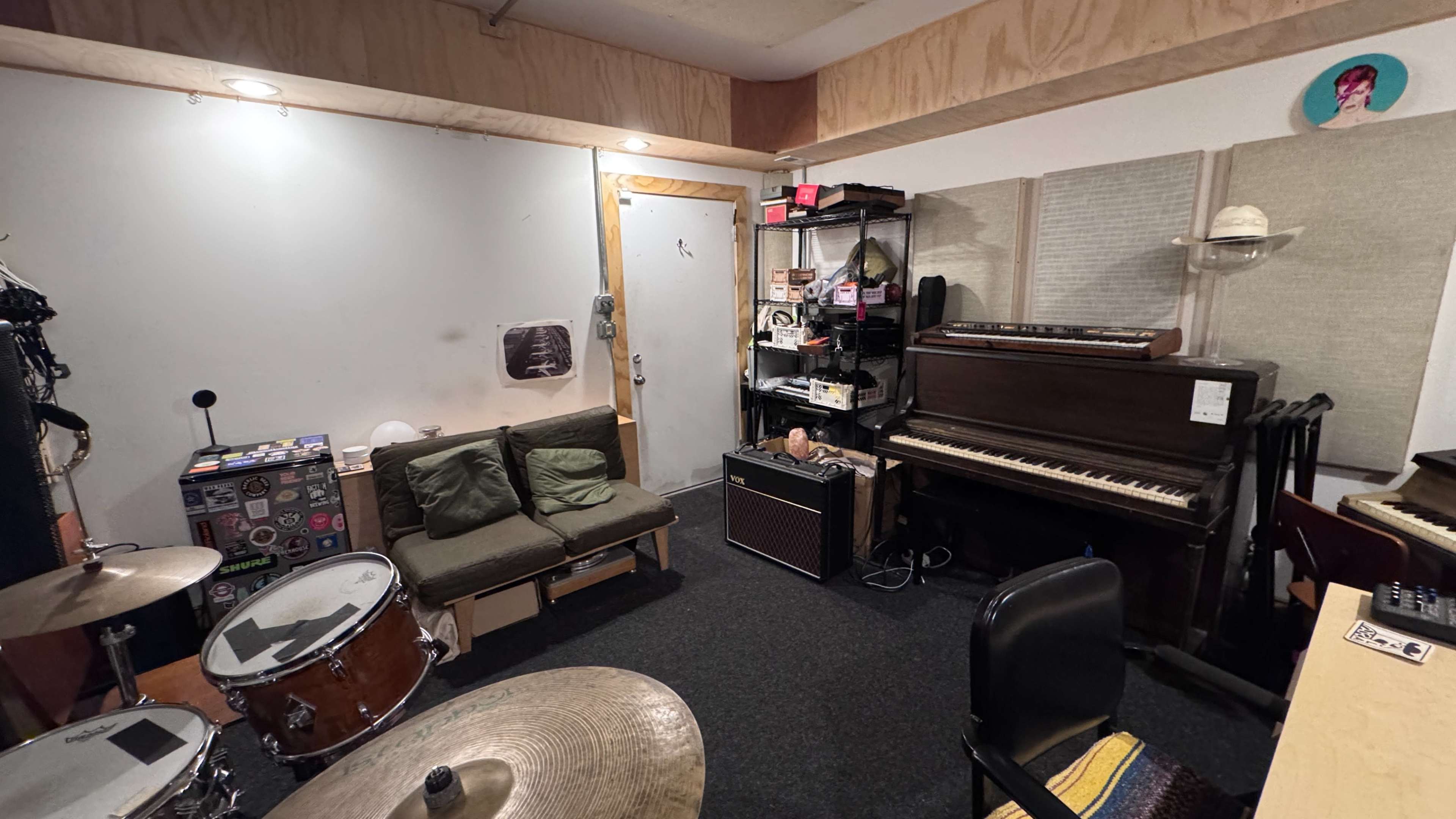 The image shows a small music rehearsal space with a drum set, a couch, a piano, and shelves filled with equipment.
