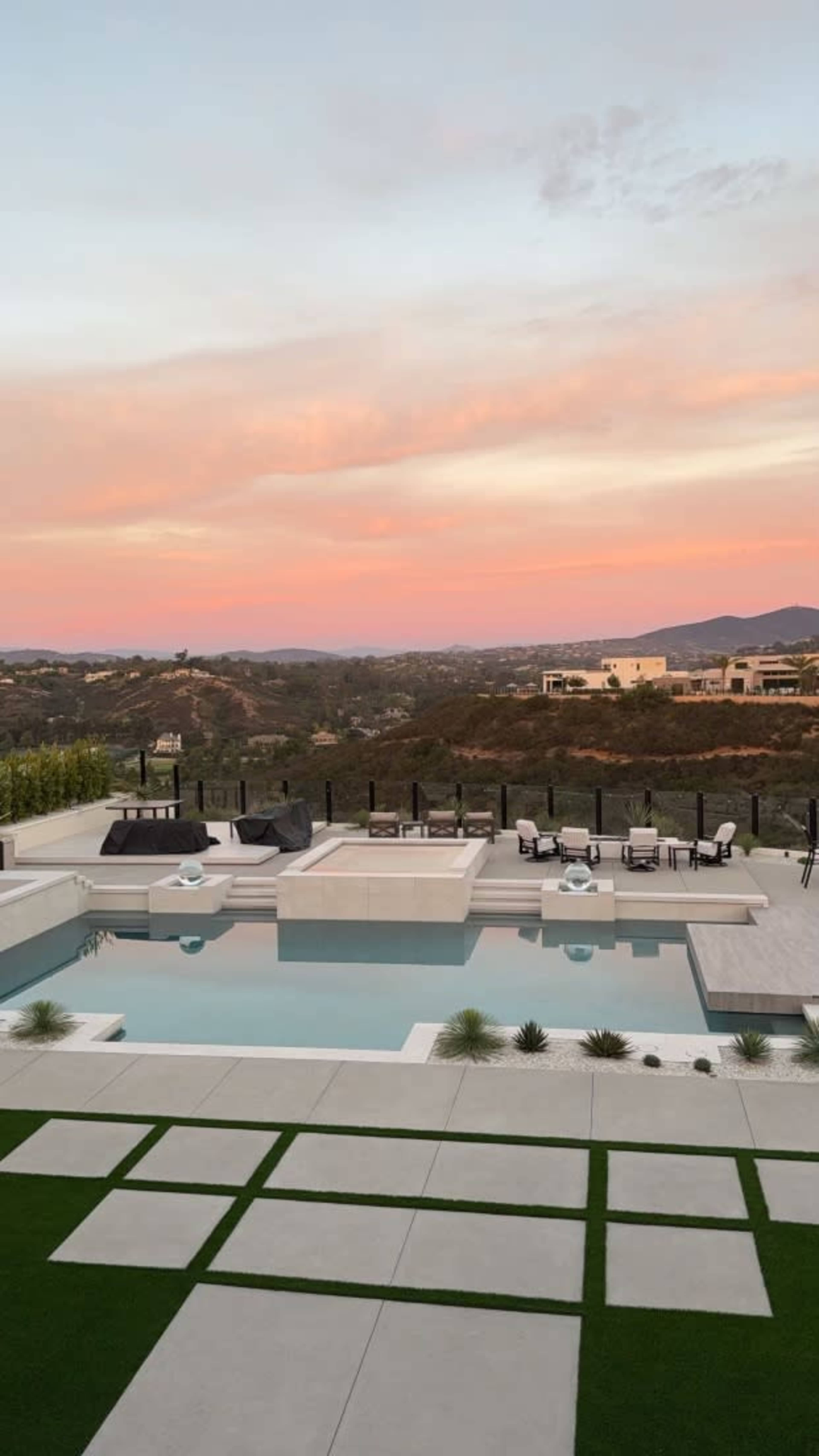 A modern outdoor area features a swimming pool, patio furniture, and landscaped greenery with a view of rolling hills at sunset.