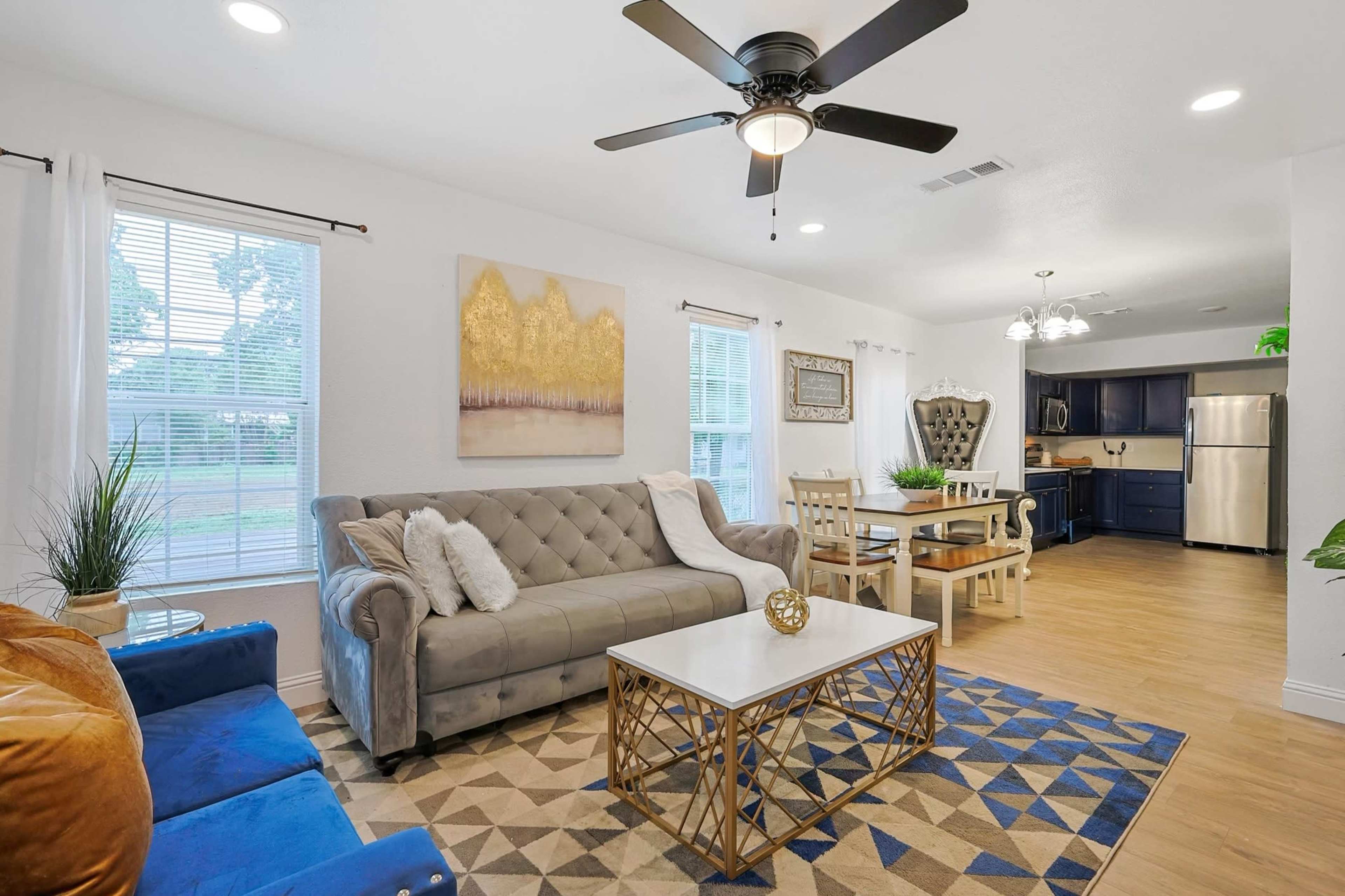 A modern living room and dining area with a gray couch, a blue armchair, a wooden dining table, and a kitchen in the background.