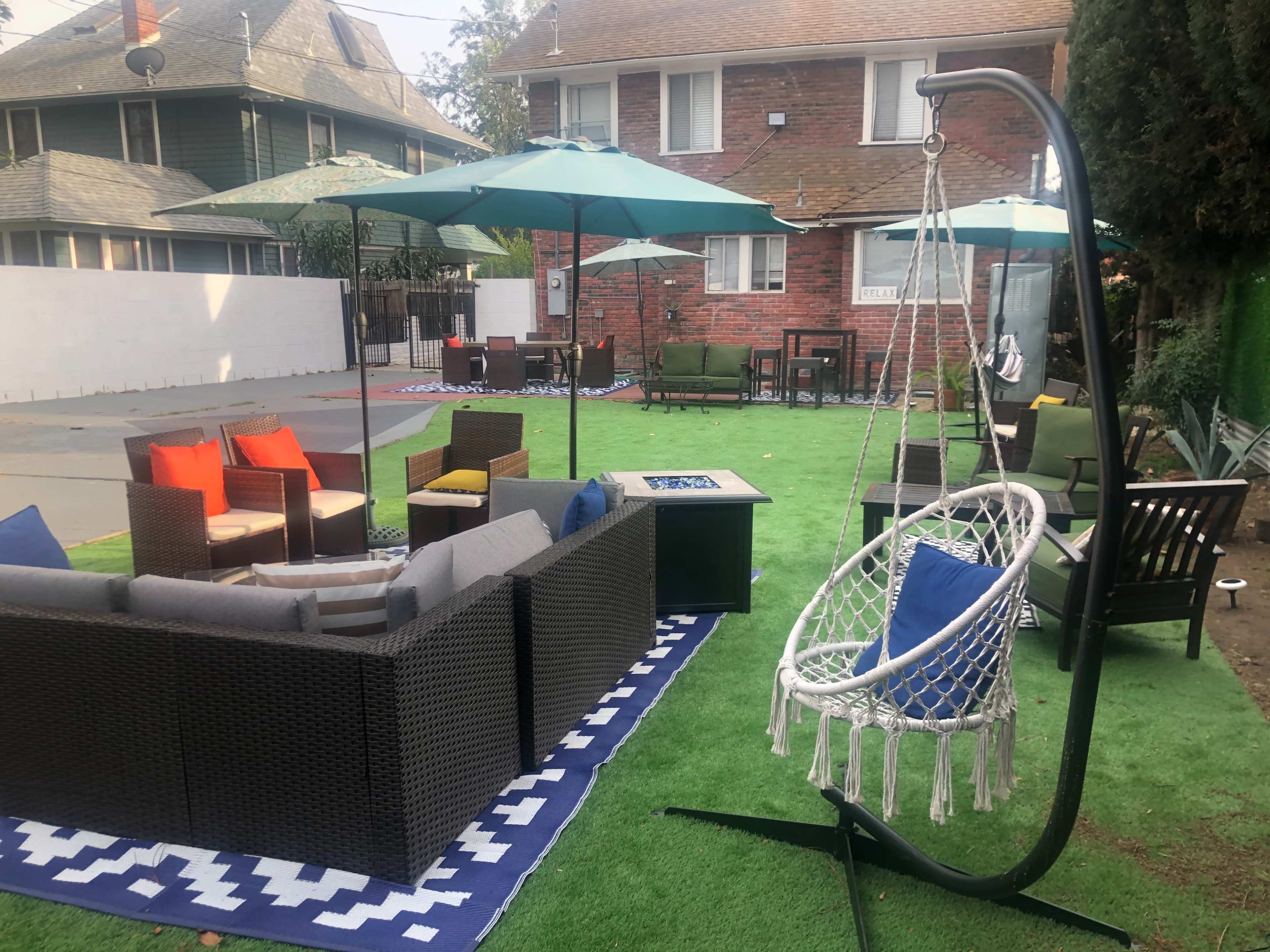 The scene features a landscaped outdoor area with seating arrangements, a swing chair, and umbrellas in a residential setting.